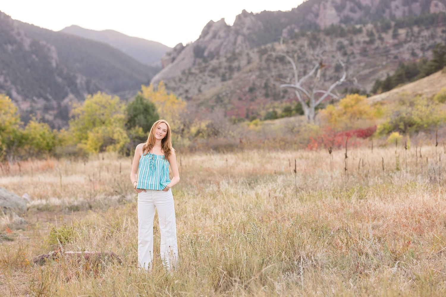 A person stands in a golden autumn meadow with rocky mountain peaks rising dramatically in the background.