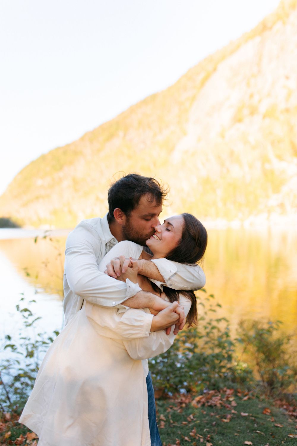 A romantic couple sharing a tender embrace against a golden sunlit wall in matching cream-colored outfits.