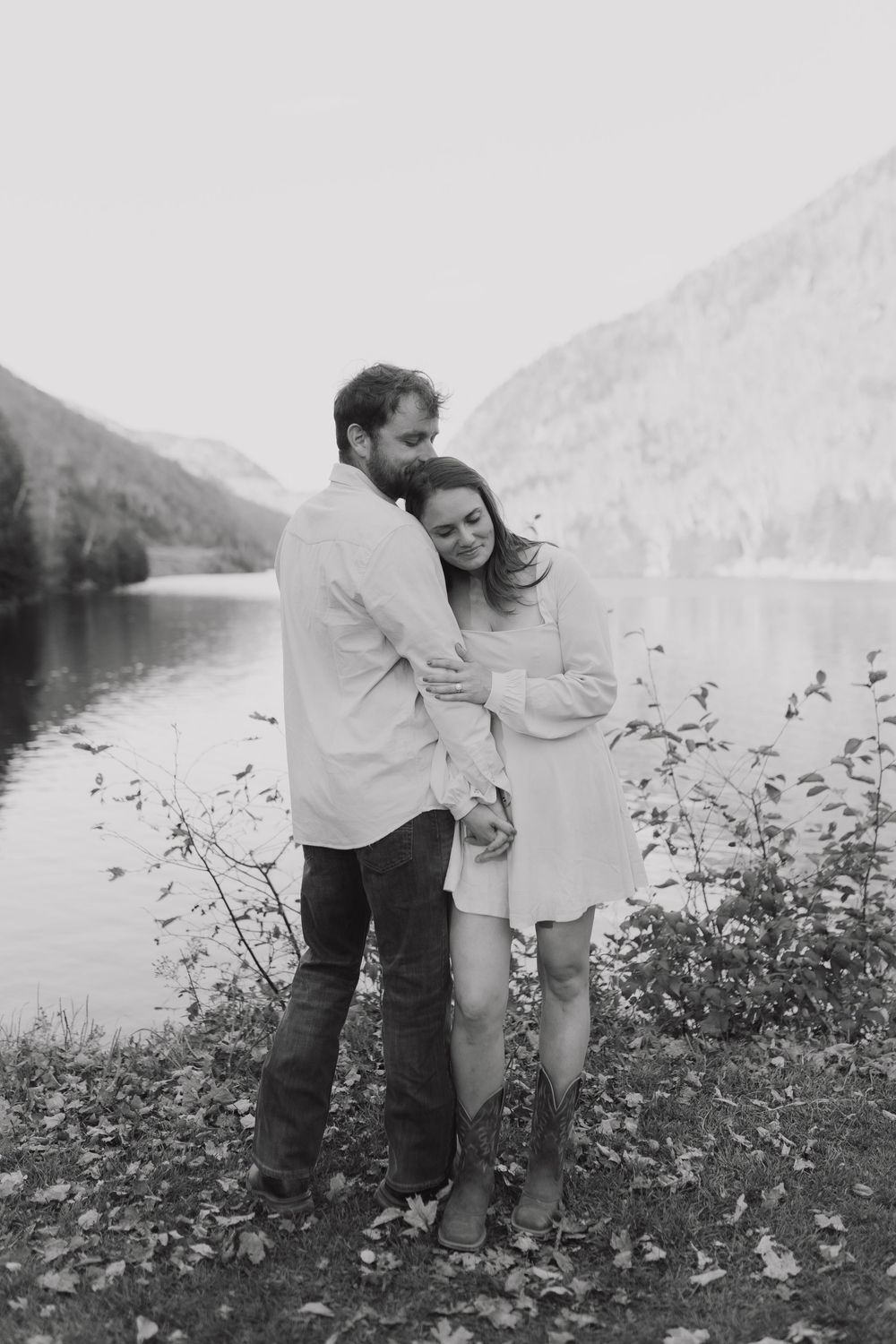 A couple in light-colored clothing picks flowers together by a serene mountain lake in a black and white photograph.