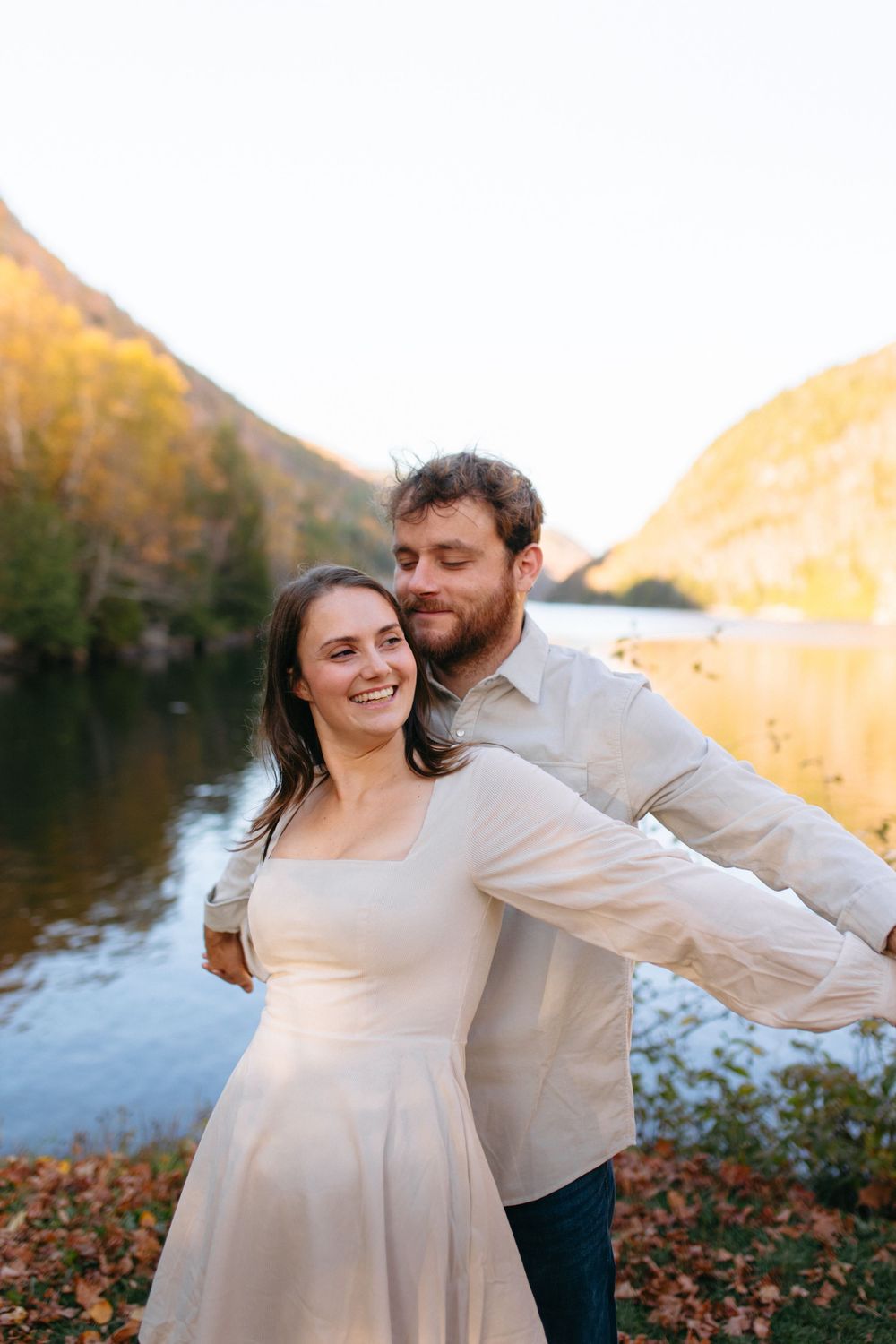 A couple in light-colored clothing shares a romantic moment by a lake during golden hour.