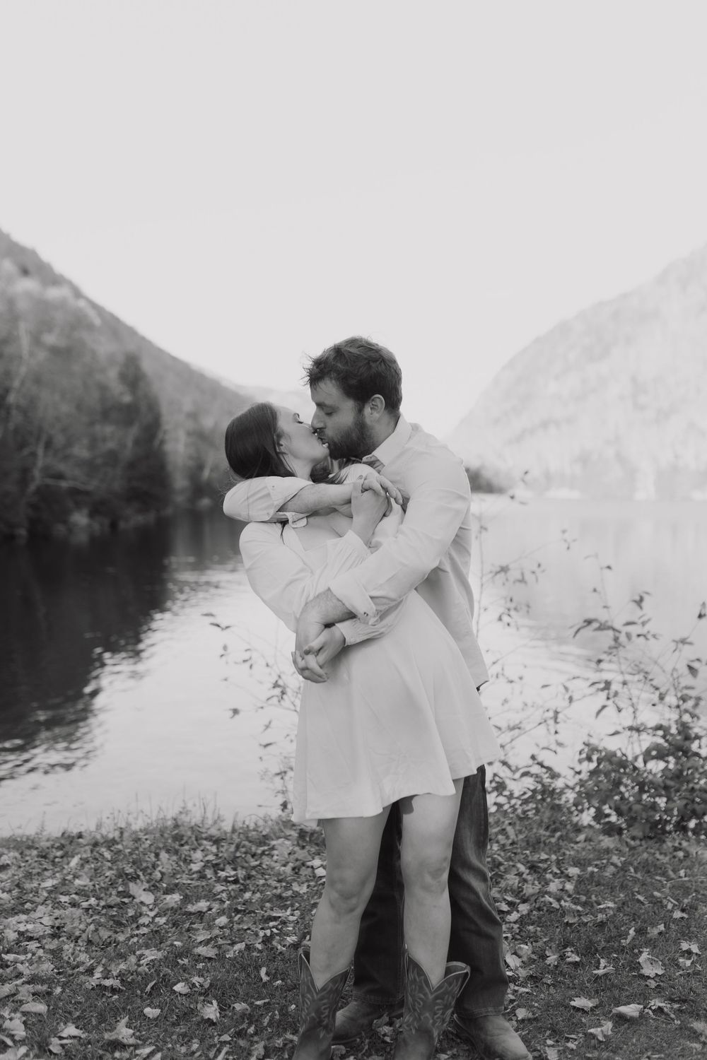 A romantic couple embraces in a field with mountains and water in the background, shown in black and white.