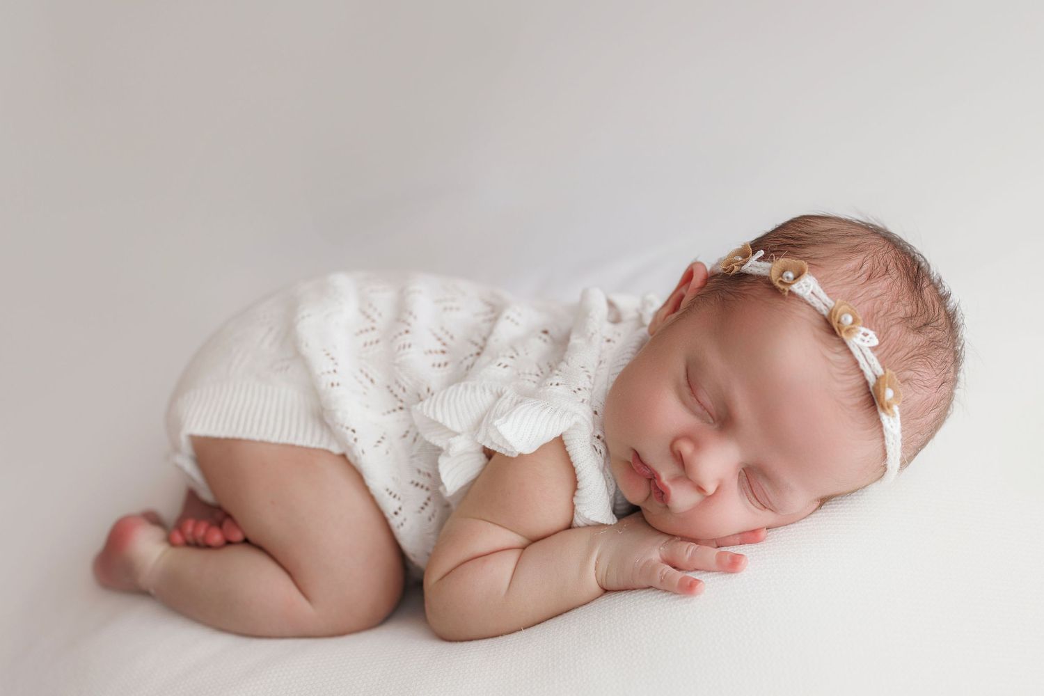 Sleeping newborn baby wearing white headband curled up in fetal position on white blanket during professional photoshoot.