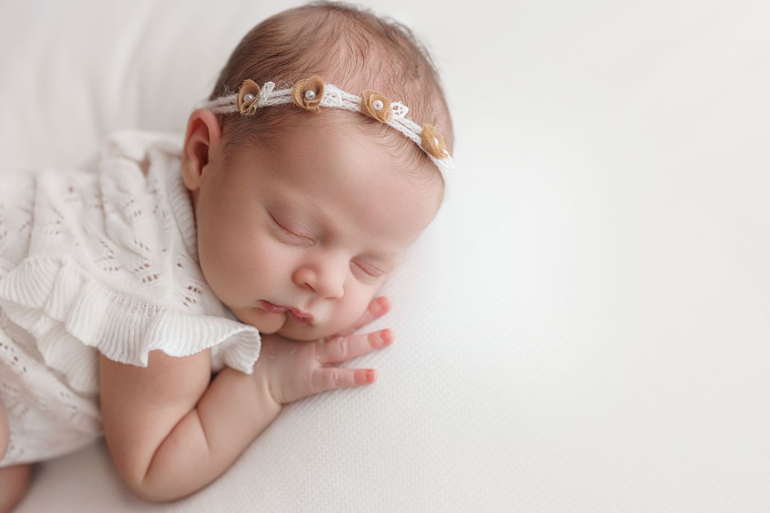 Sleeping newborn in white lace dress and delicate headband rests peacefully on soft white backdrop.