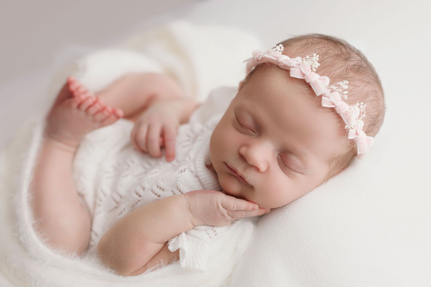 Sleeping newborn baby in white lace headband resting peacefully on soft white bedding.