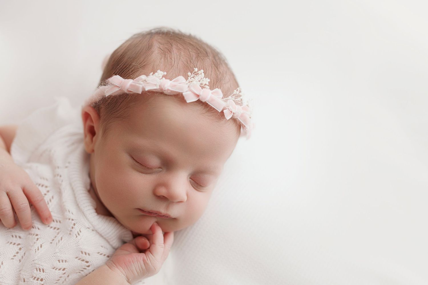 Sleeping newborn wearing delicate white lace dress and pink headband rests peacefully on soft white blanket.