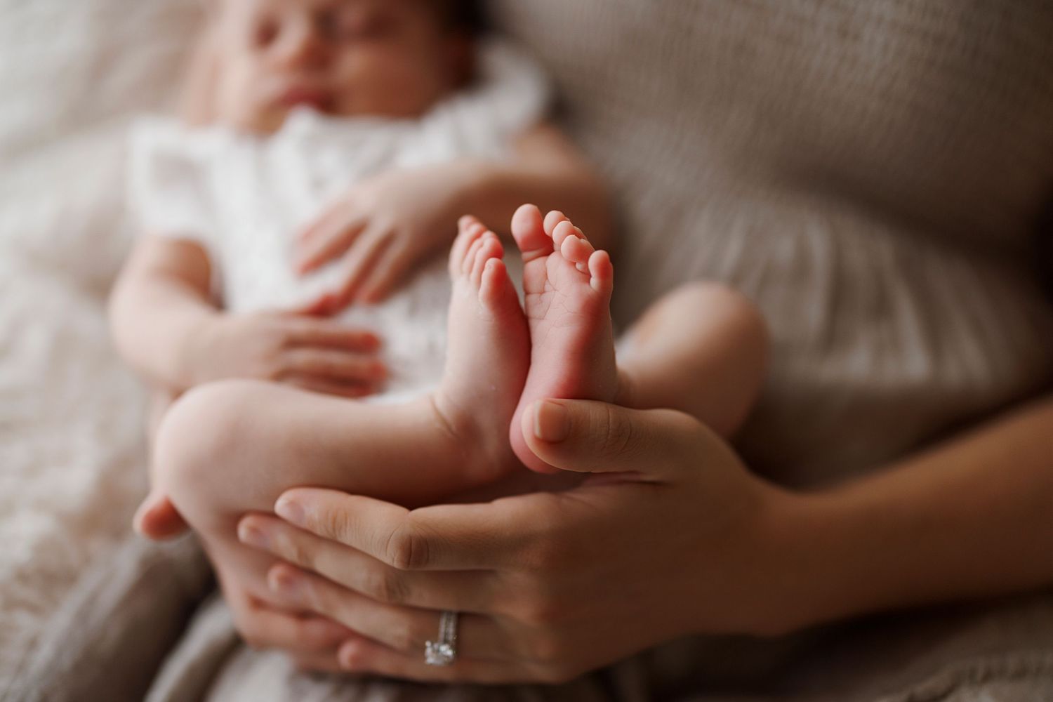 Close-up of tiny baby feet being gently held by adult hands with engagement ring on soft bedding.