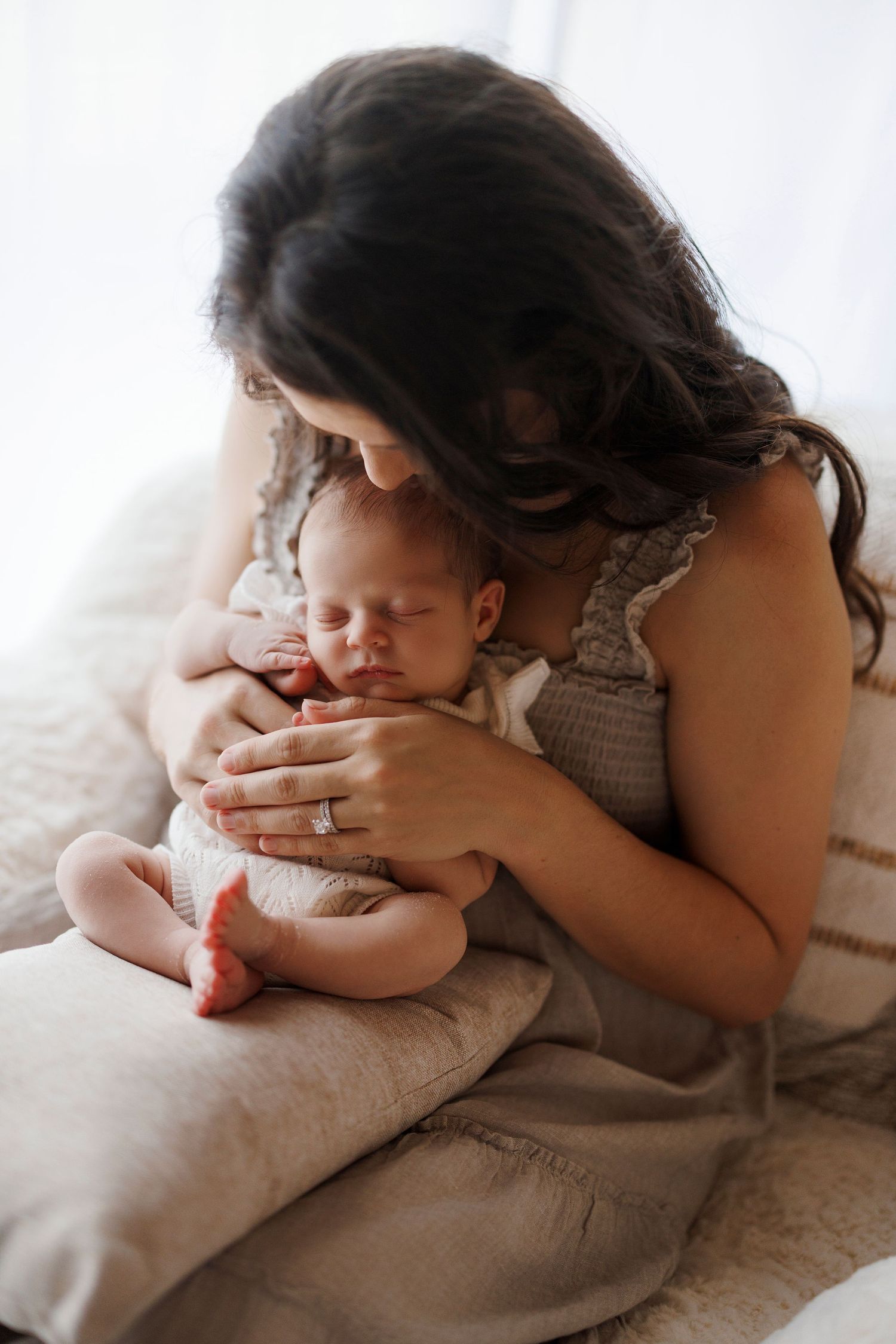 An intimate portrait of a parent in a beige dress holding and kissing a sleeping newborn baby on a plush chair.