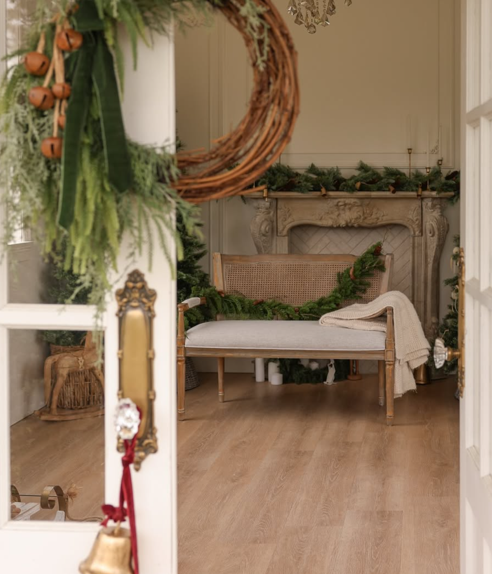 Rustic holiday interior with grapevine wreath, evergreen garland, and wooden accents against cream colored walls.