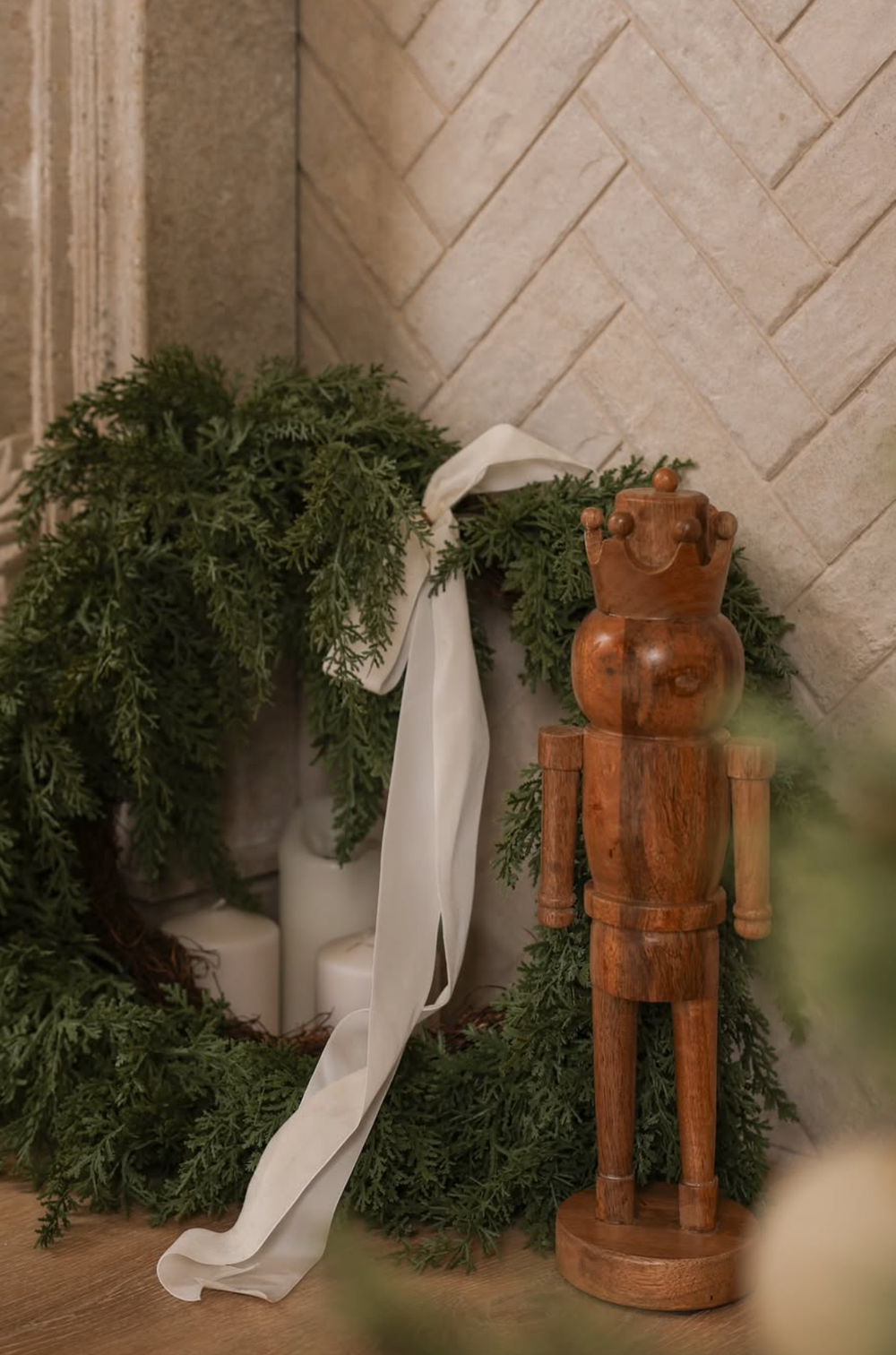 Rustic wooden nutcracker figure stands beside evergreen garland and white ribbon against a textured wall.