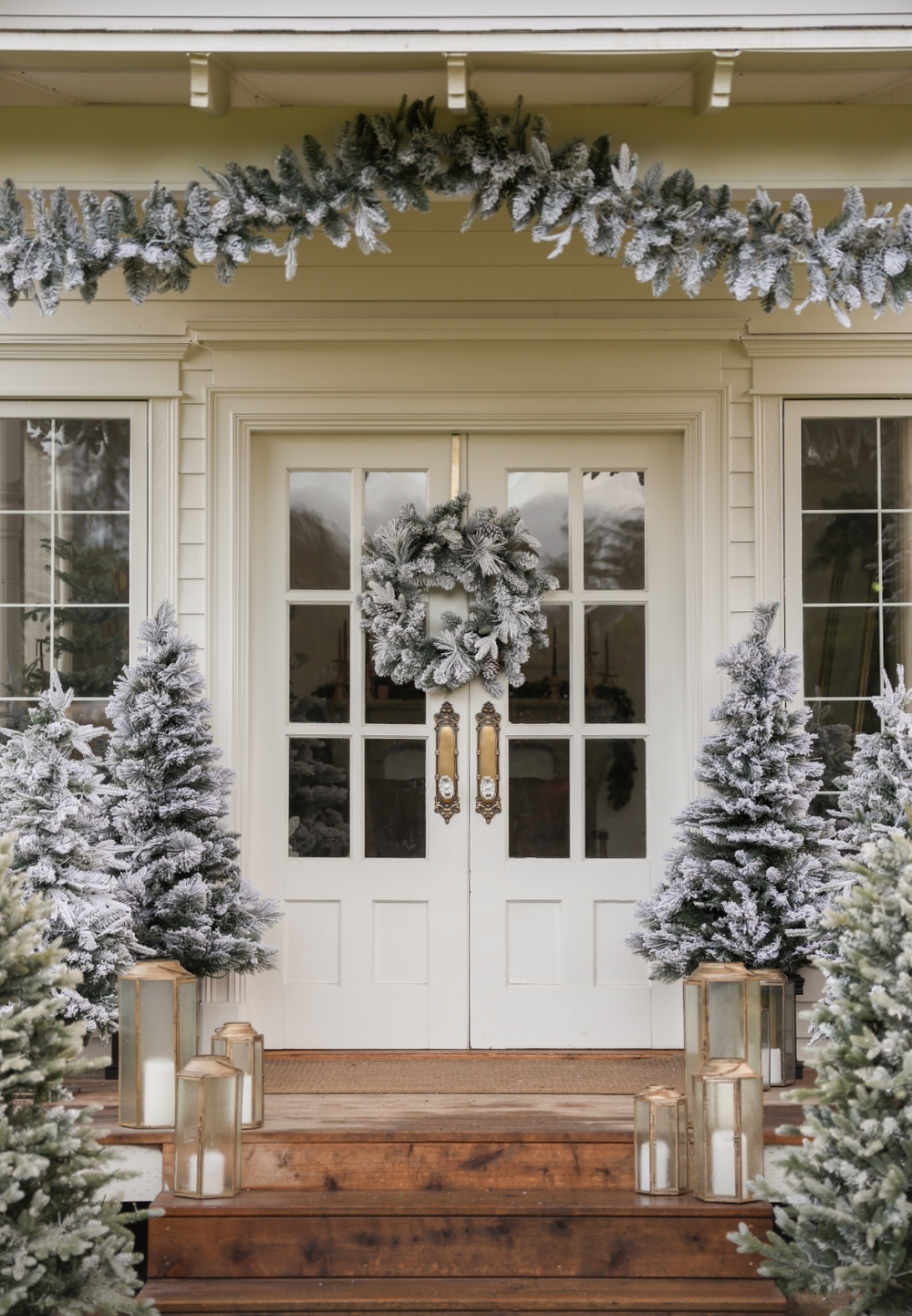 Snow-covered evergreen trees flank a white wooden entrance with double doors and wooden benches on the porch.