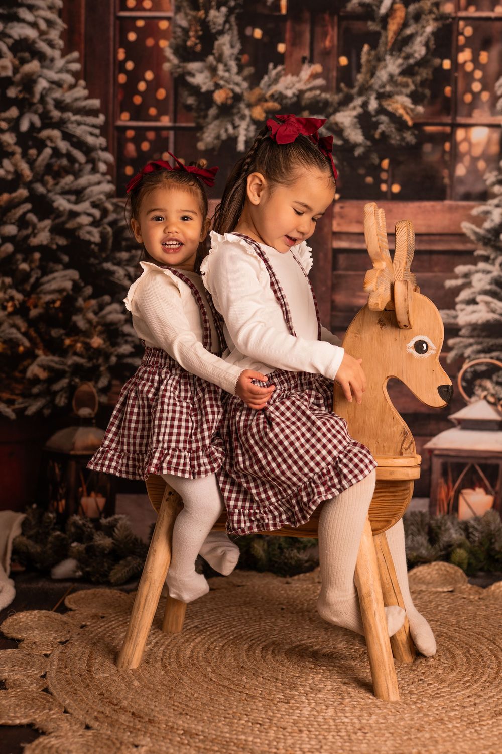 Two children in matching plaid dresses and white tights pose on a wooden rocking reindeer with Christmas decor in background.