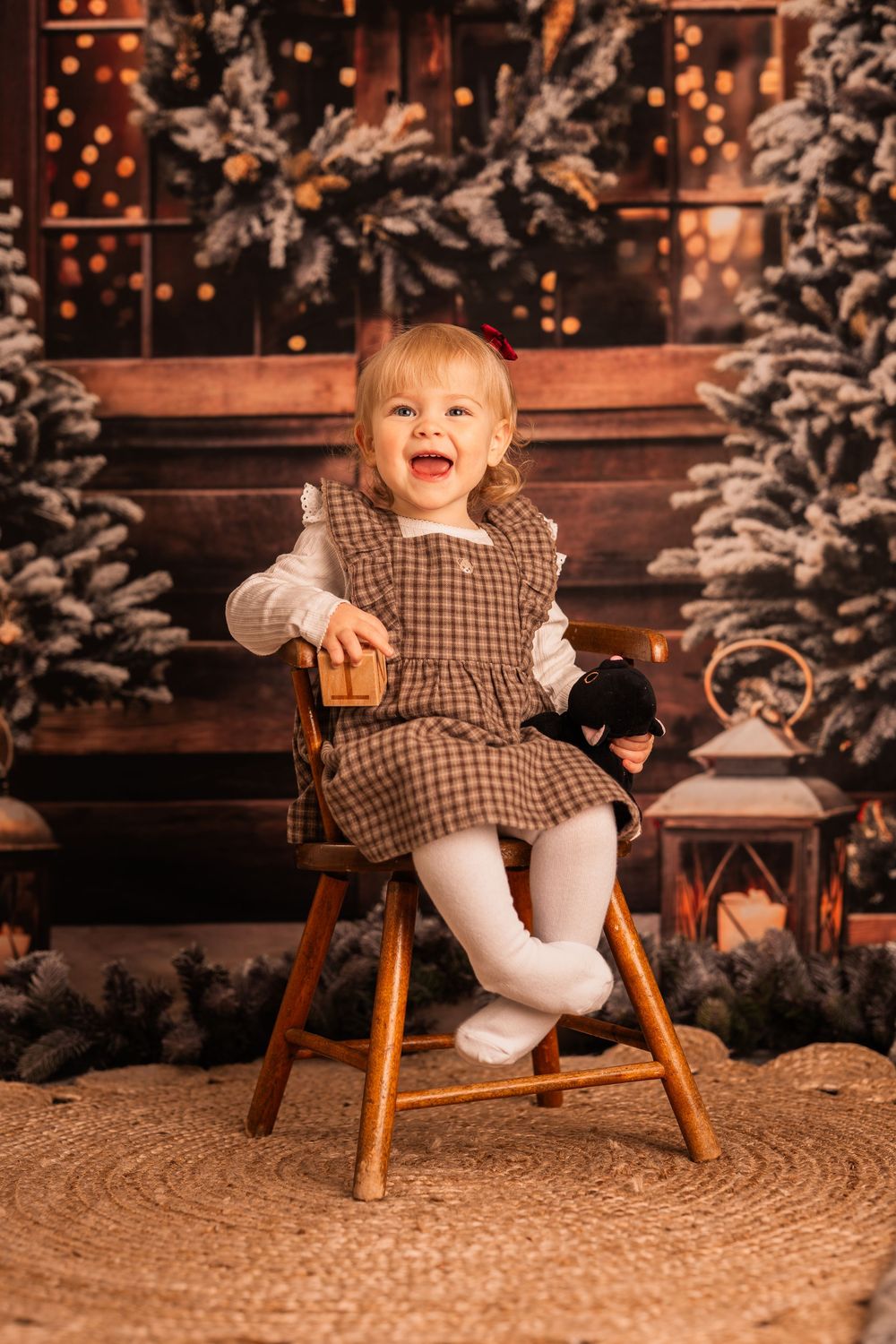 A small child in a plaid dress and white tights sits on a wooden stool against a festive holiday backdrop with bokeh lights.