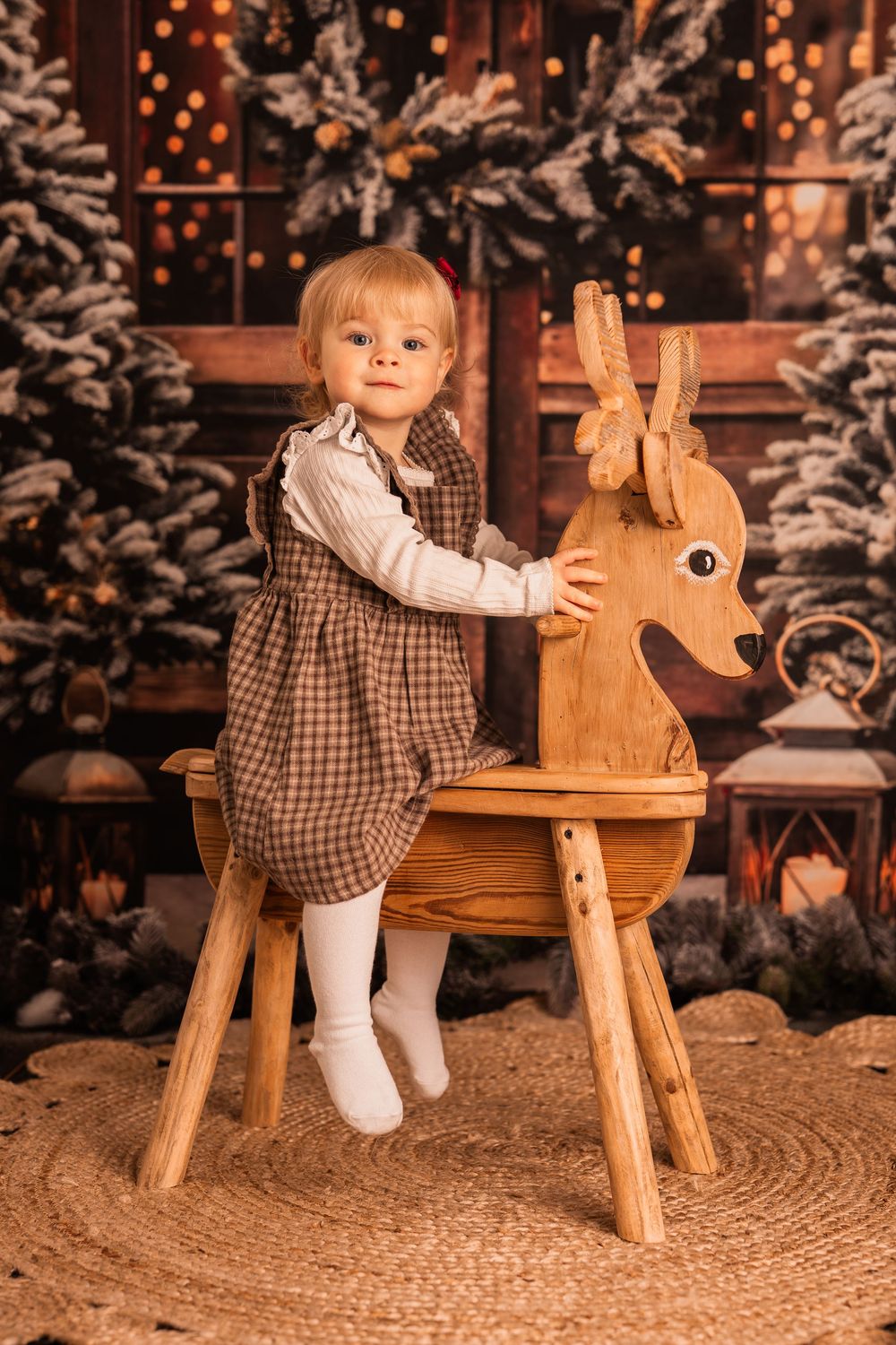 A small child in a checkered dress plays with a wooden rocking horse in a cozy Christmas setting with snowy trees.
