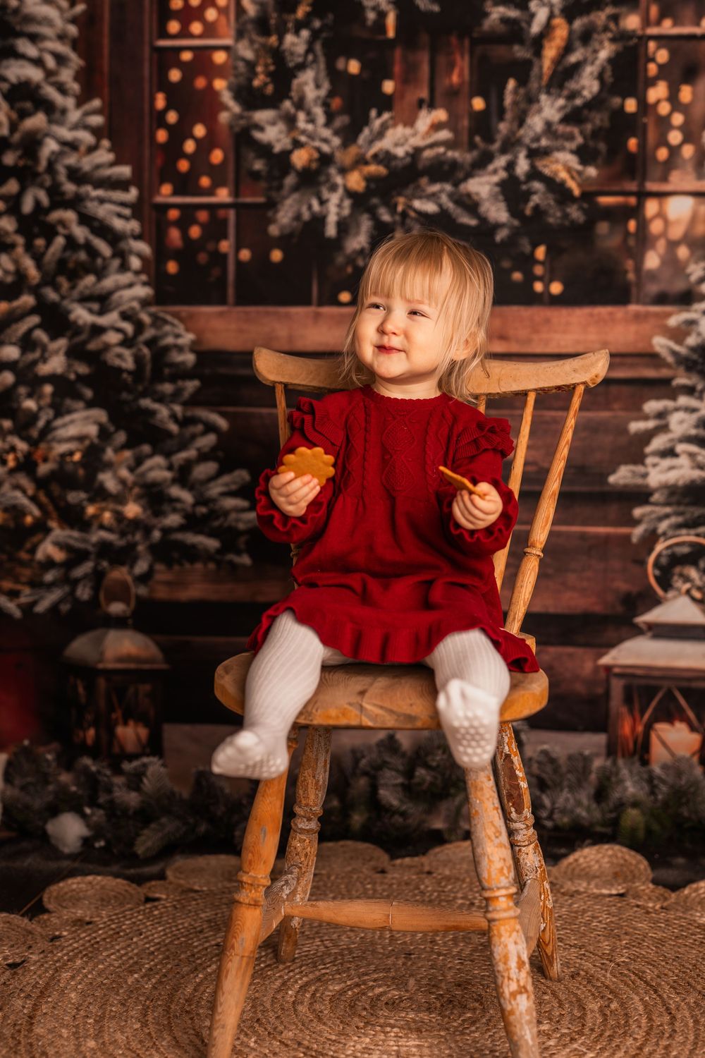 Small child in red sweater and white tights sits on wooden chair surrounded by snowy Christmas decorations.