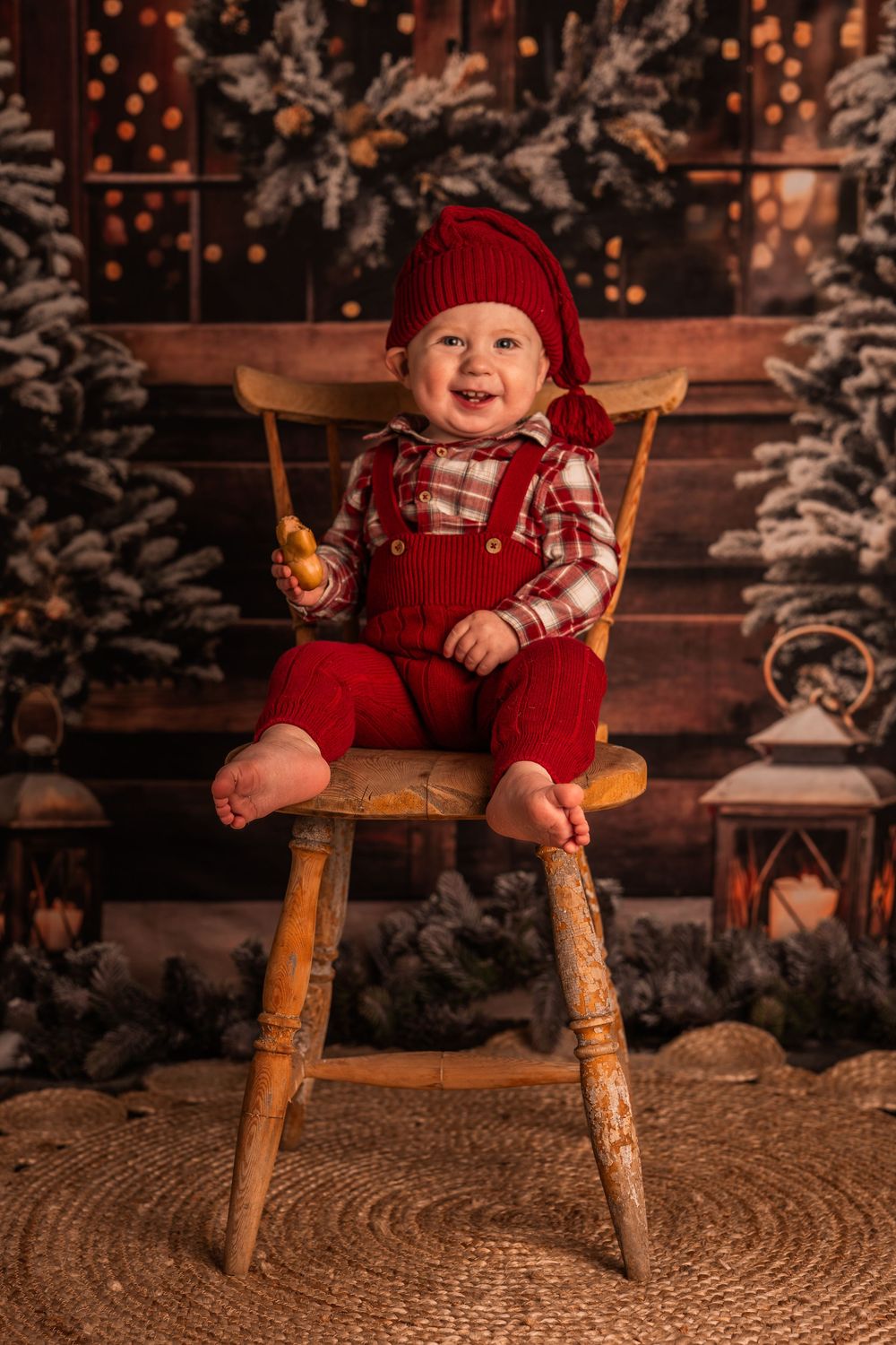 Baby in red overalls and knit hat sits on wooden chair with snowy Christmas scene and twinkling lights in background.