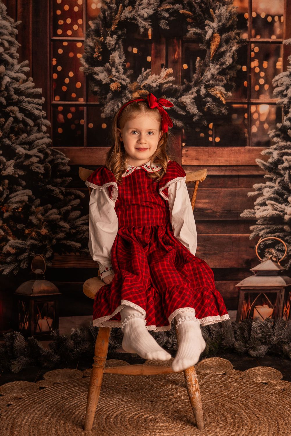 Young girl in red plaid Christmas dress sits on wooden stool surrounded by snow-flocked trees and lanterns.