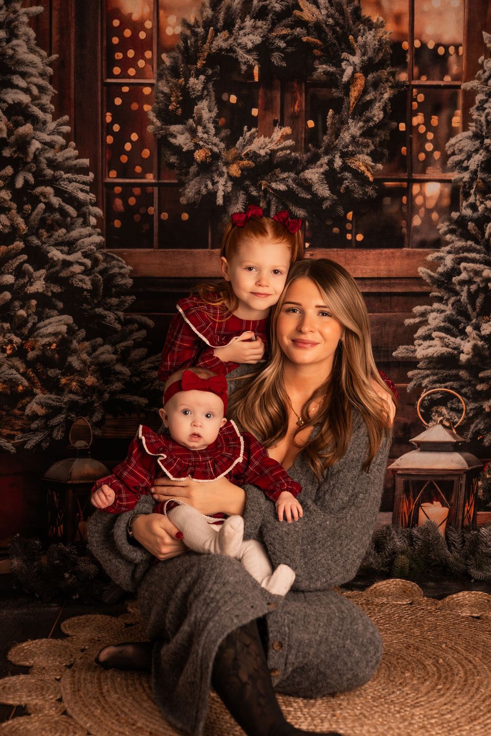 A mother sits with two young children in red plaid outfits among snowy Christmas trees and warm bokeh lights.