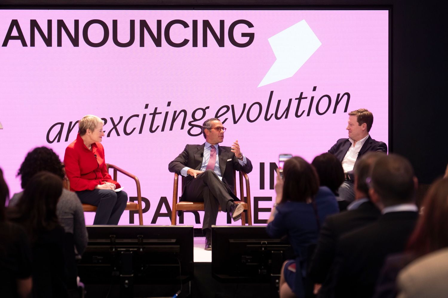 People seated in an auditorium with pink lighting and projection screens displaying text during an event presentation.
