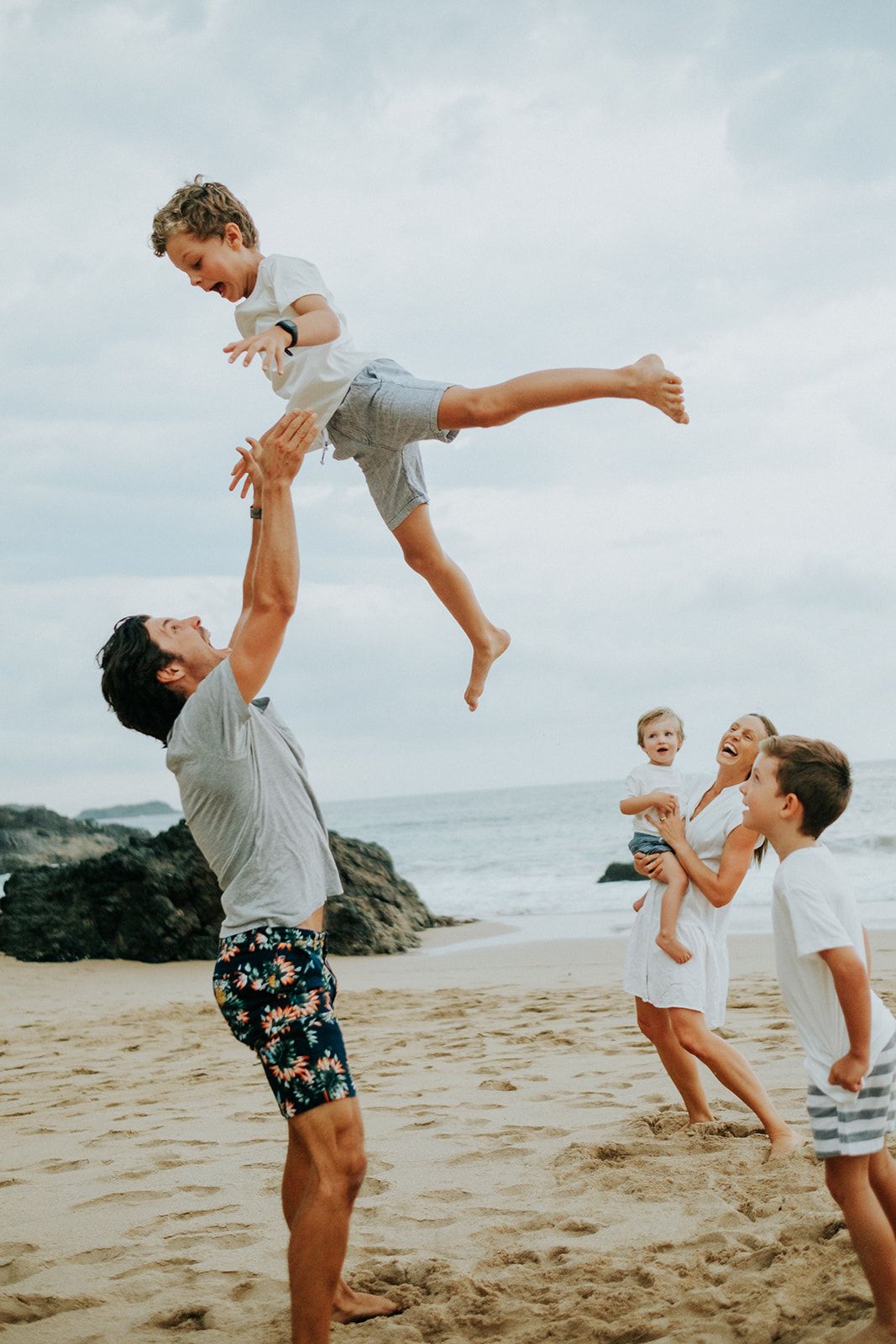 Playful beach scene with people tossing children in the air against cloudy sky.