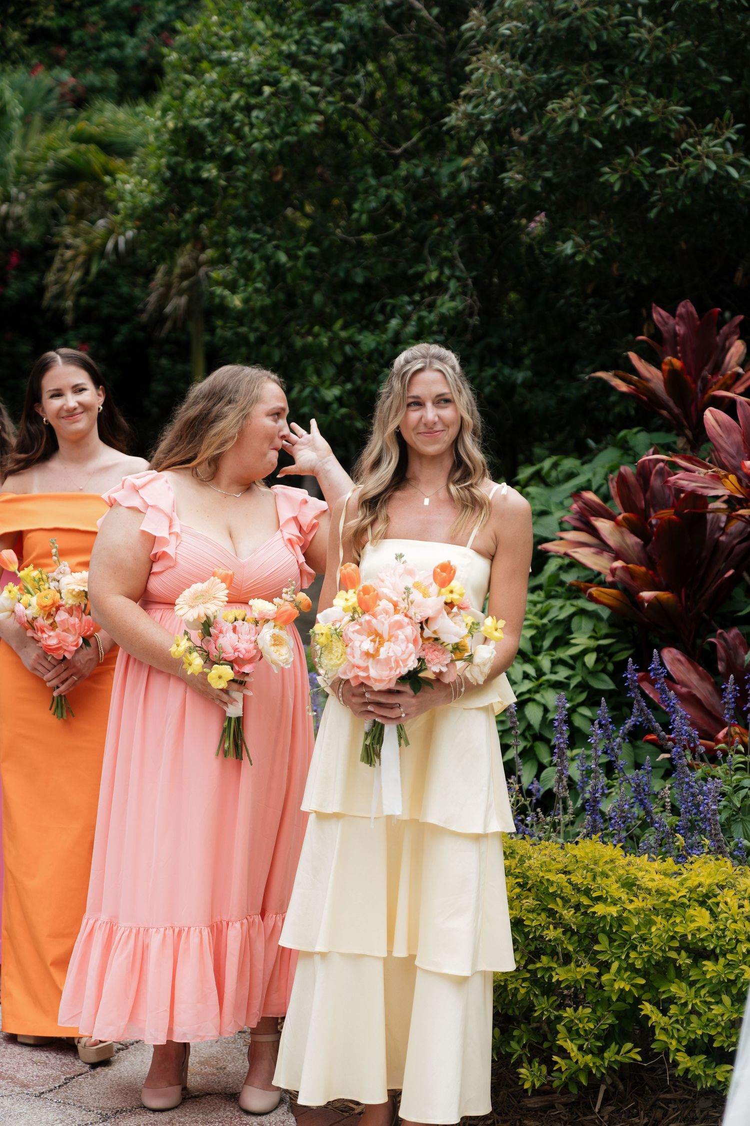 Bridesmaids in coral and cream dresses hold matching floral bouquets while standing in a lush garden setting.