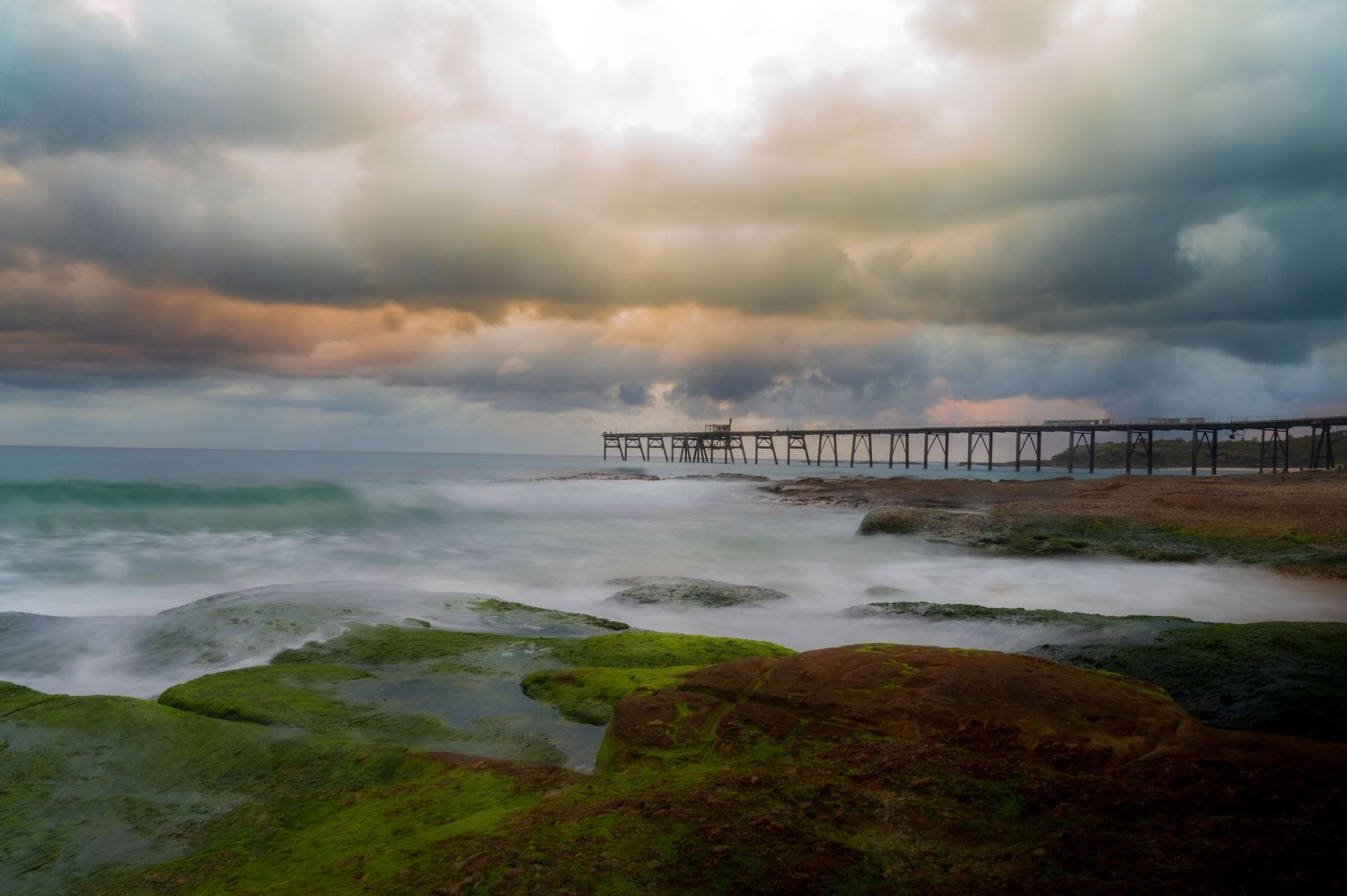 Dramatic coastal landscape with storm clouds gathering over a distant pier at dusk.