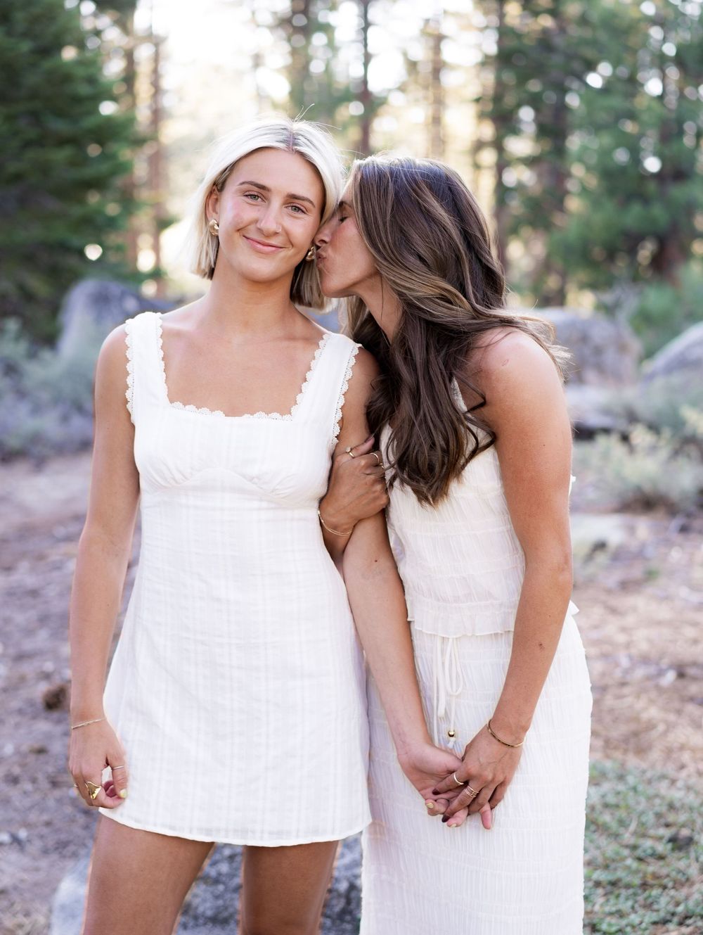 Two people in white dresses share joyful moments together in a forest setting during golden hour.
