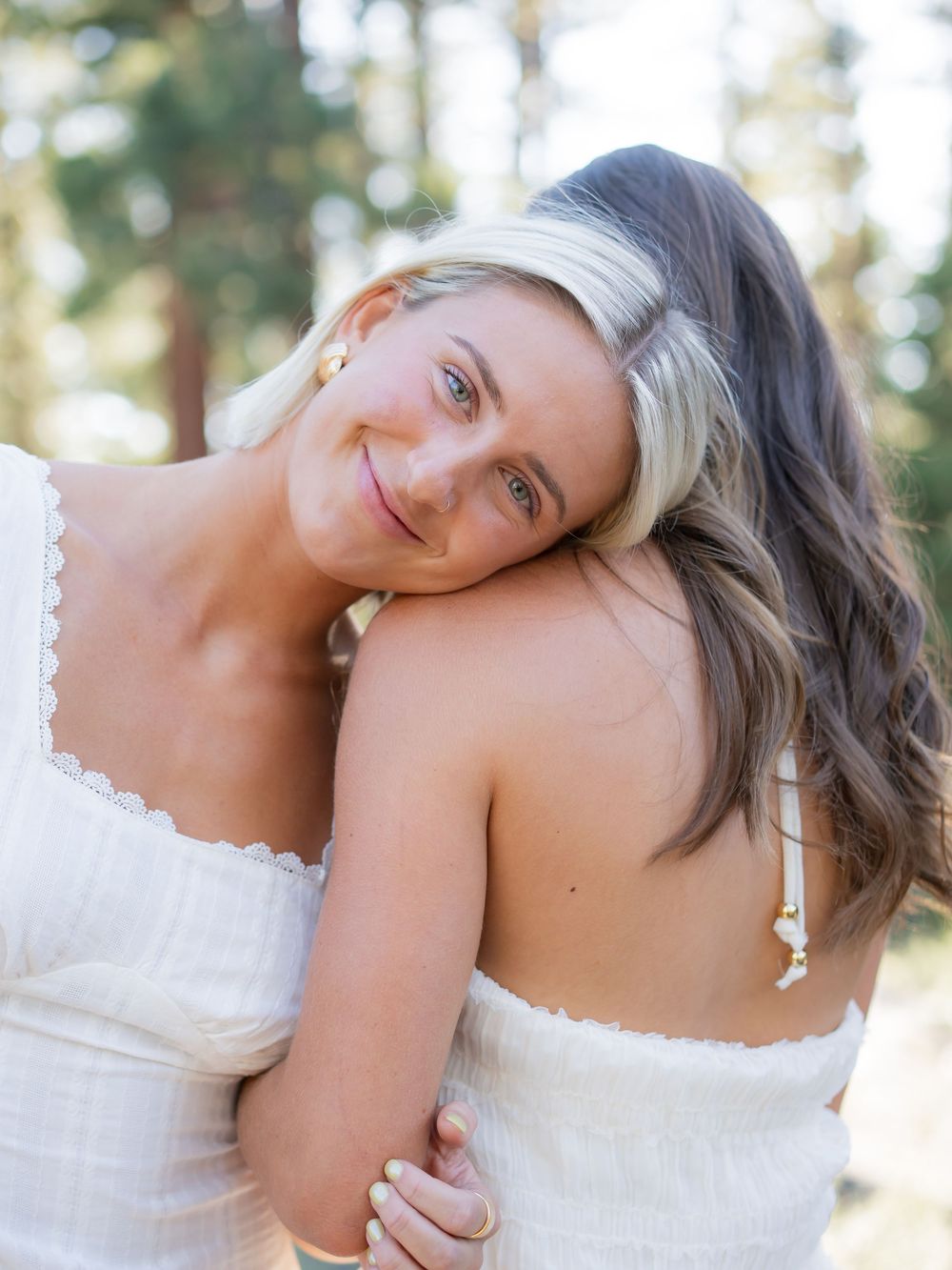 Two people in white wedding attire embrace tenderly against a sunlit forest backdrop.