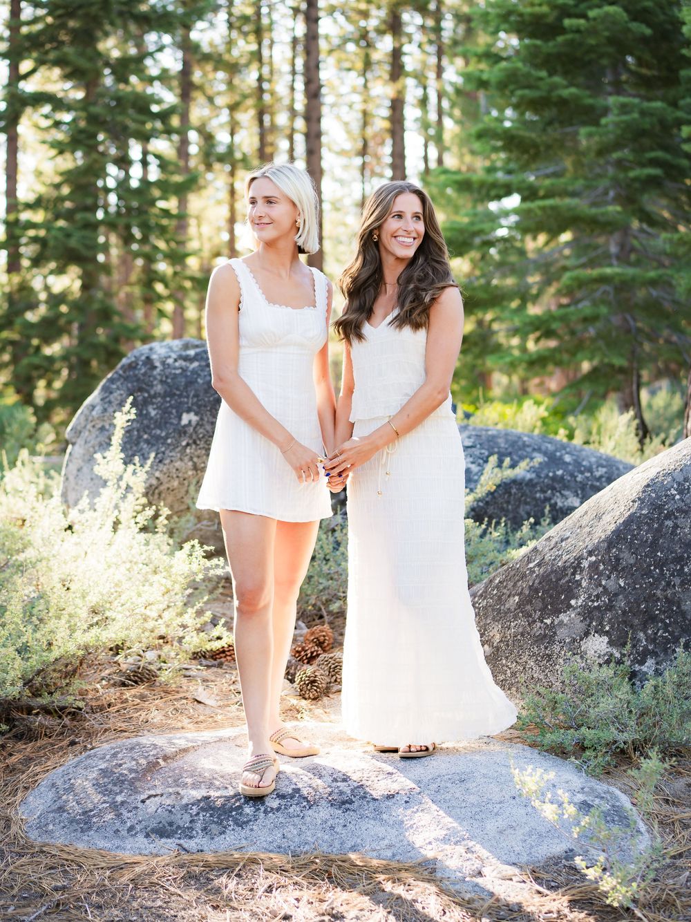 Two people in white dresses pose together on rocky terrain surrounded by pine trees in golden sunlight.