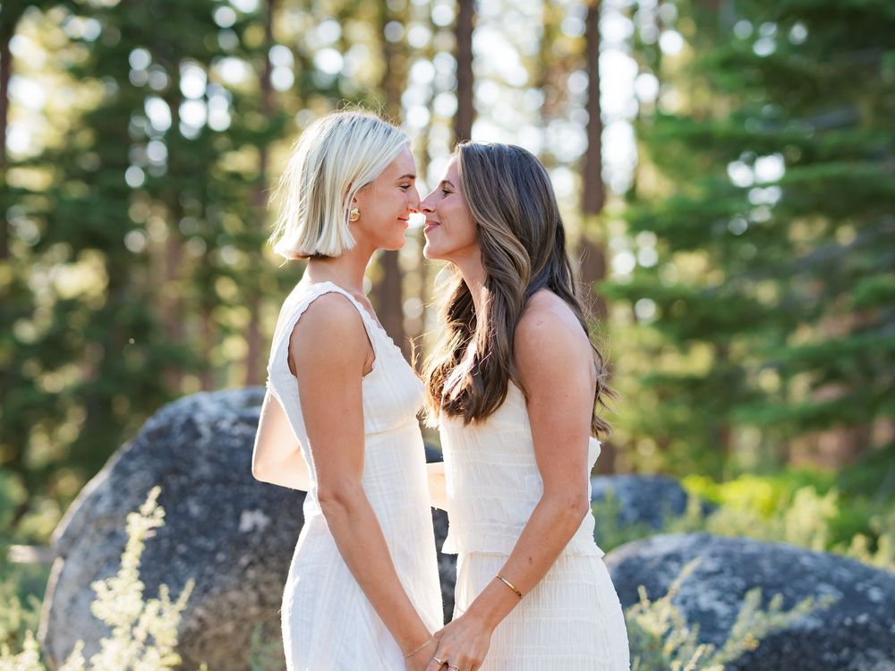 Two people share an intimate moment while standing in a forest setting with sunlight filtering through the trees.