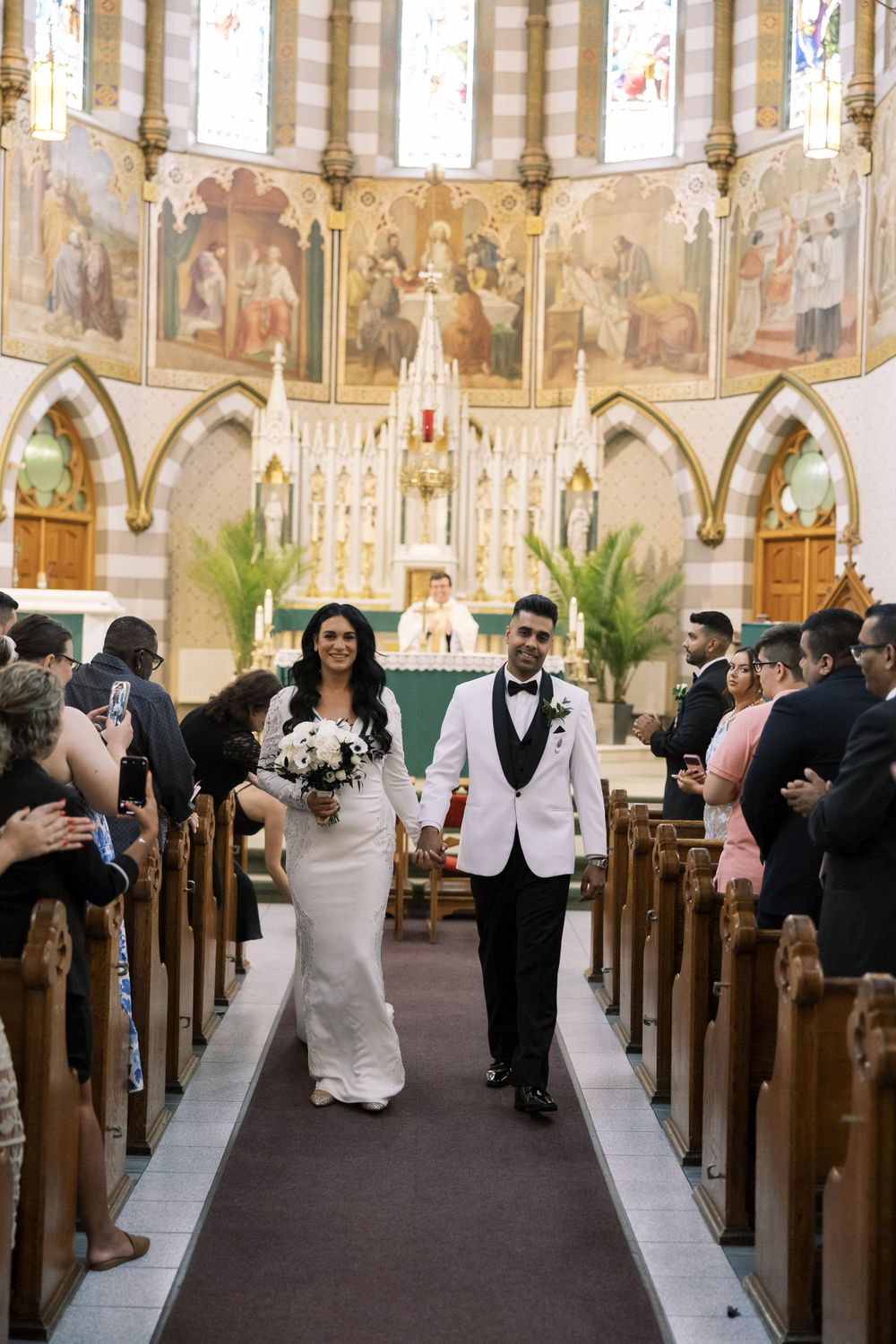 A wedding couple walks down the aisle in a beautiful church with ornate religious murals and gothic architecture.