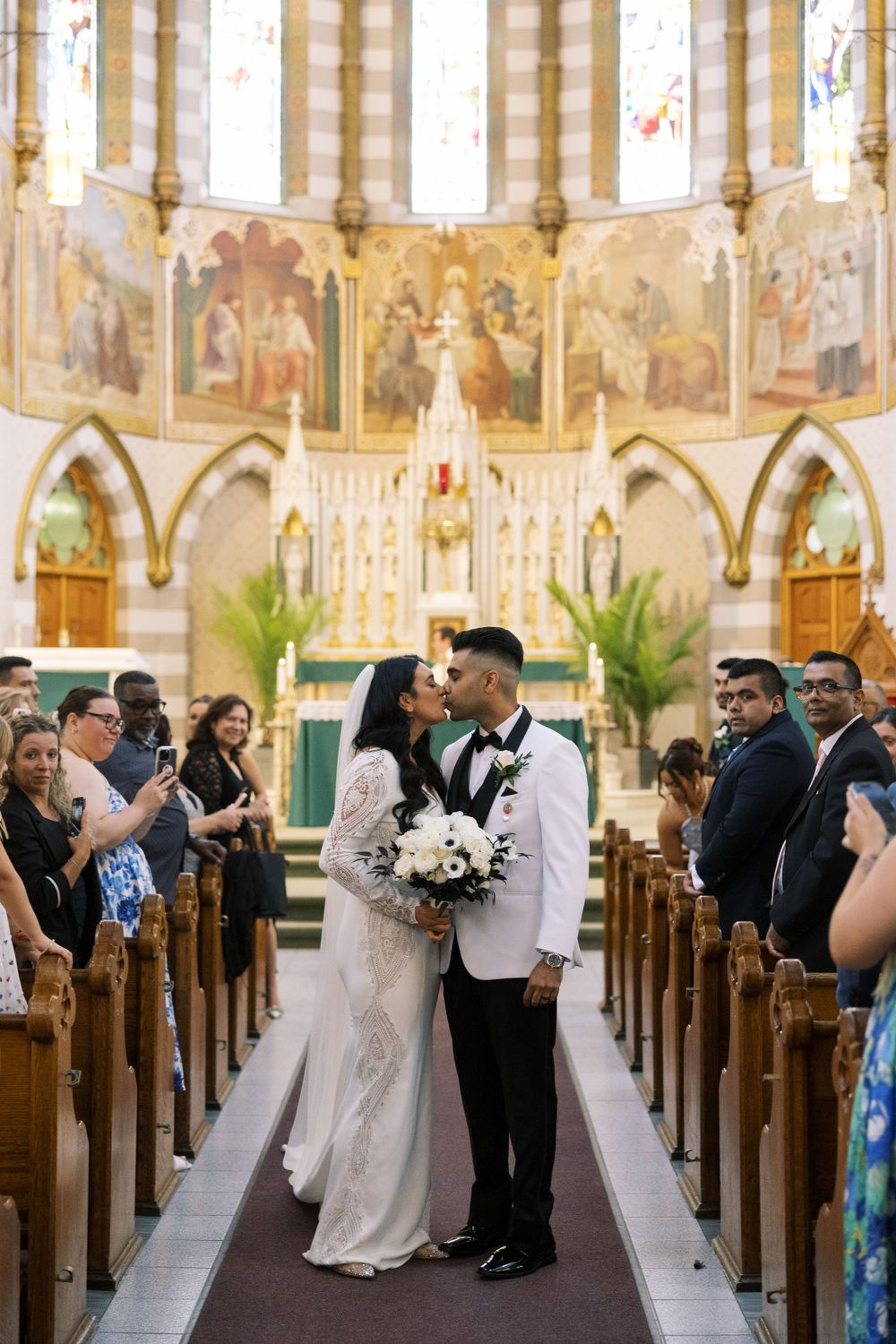 A wedding ceremony takes place in a beautiful historic church with ornate stained glass windows and religious artwork.