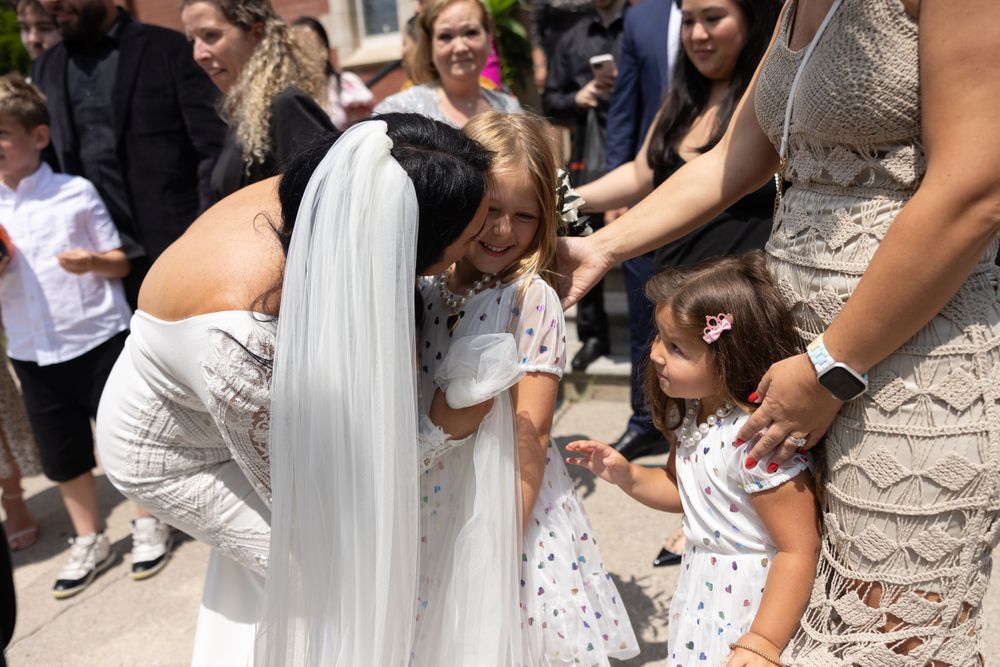 A bride in a white wedding dress bends down to hug two children in matching white dresses at an outdoor event.
