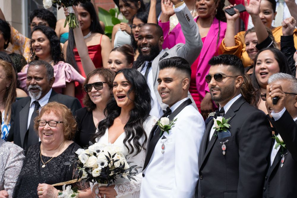 A joyous wedding party poses together for a group photo, with people in formal attire and bright smiles.