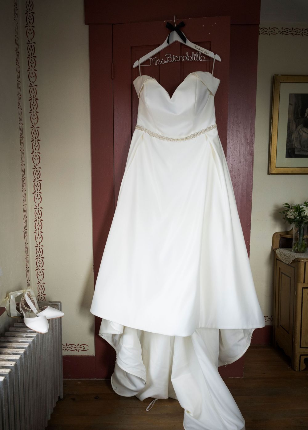 An elegant white wedding dress with sweetheart neckline hangs on display against a burgundy wall.