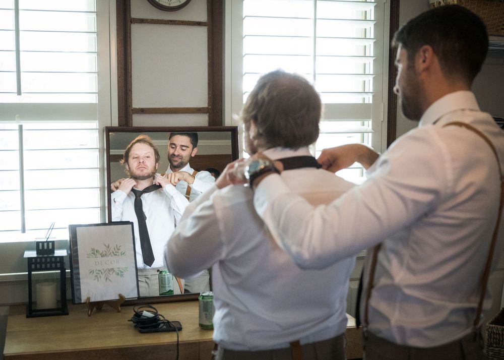 Two people adjust their bow ties while getting ready in front of a mirror with window light.
