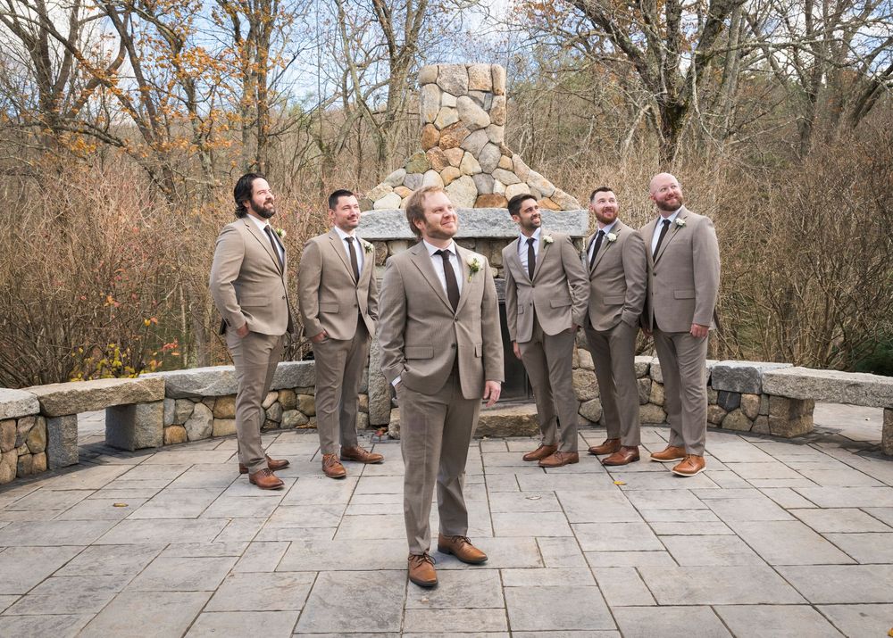 A wedding party in matching gray suits poses playfully on a stone patio surrounded by winter trees.