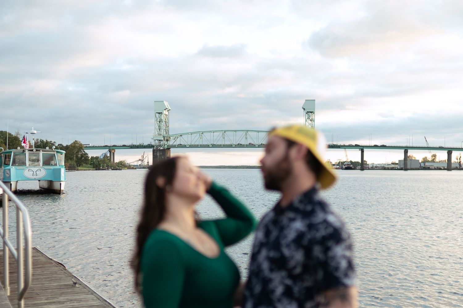 Two people embrace and share a romantic moment on a waterfront pier with a bridge visible in the background.