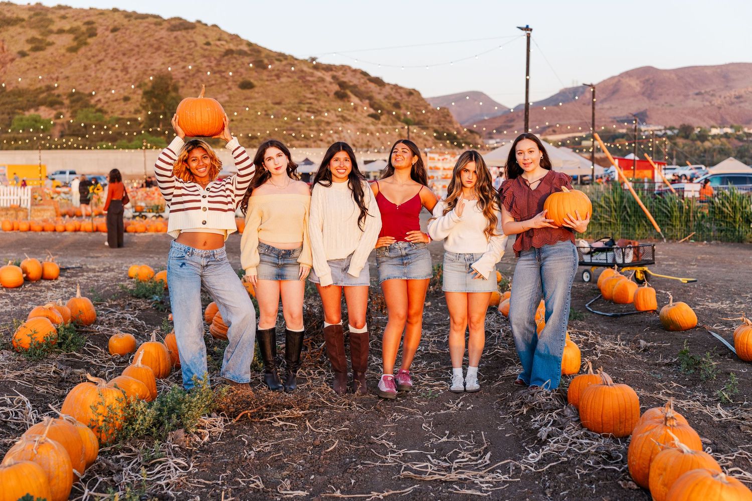 Group sits among bright orange pumpkins at sunset in a rustic pumpkin patch setting.