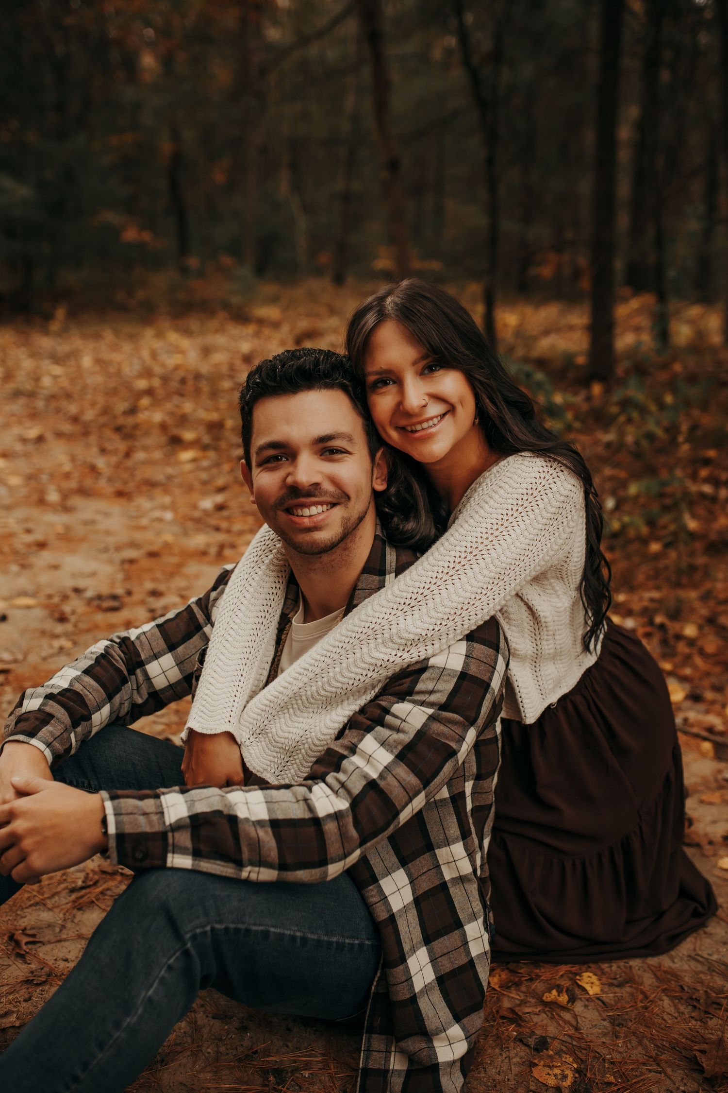 A couple sits together on fallen autumn leaves wearing cozy sweaters and plaid in a romantic outdoor setting.
