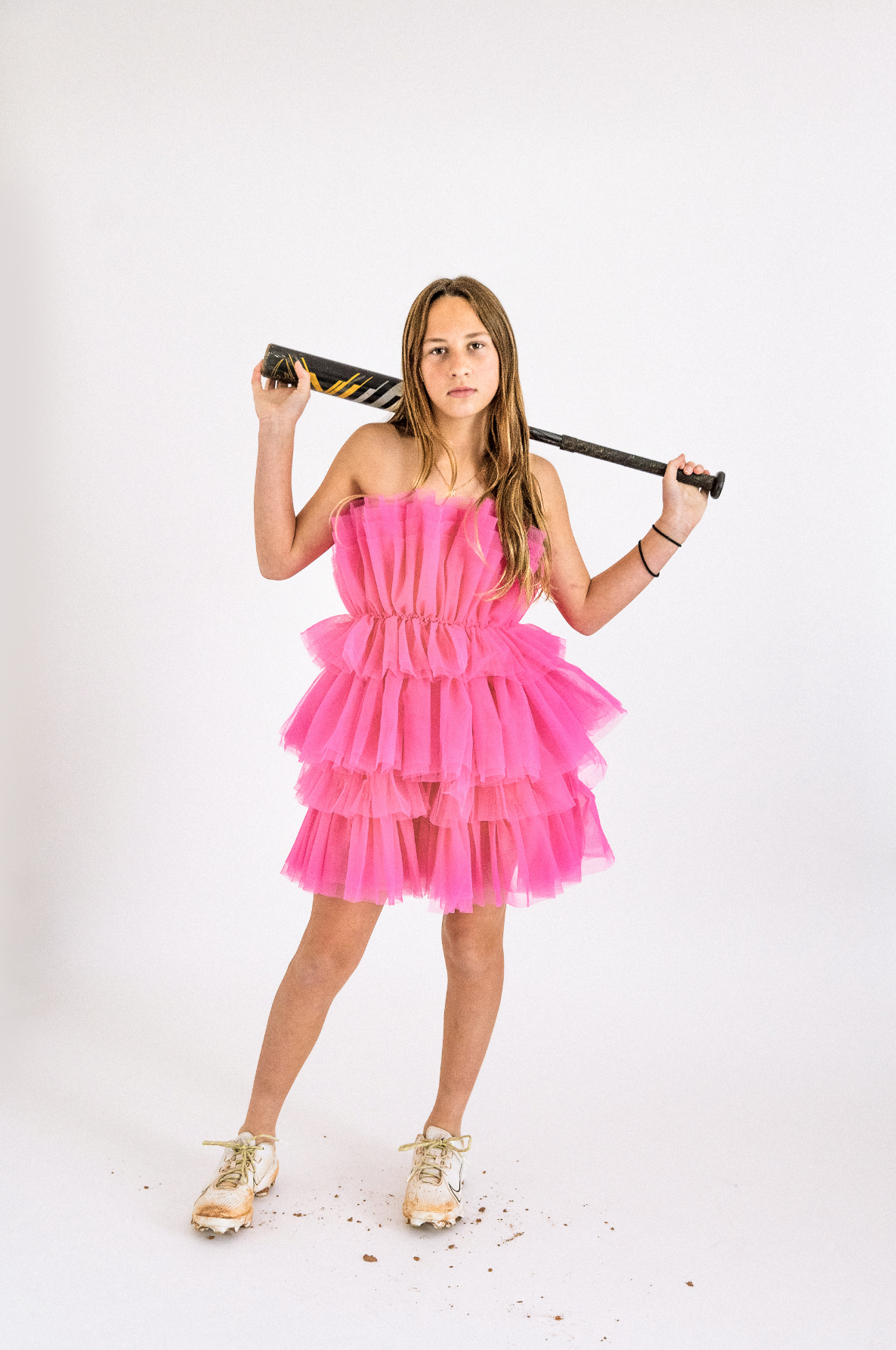 Person in pink ruffled dress poses with baseball bat against white background while wearing gold shoes.