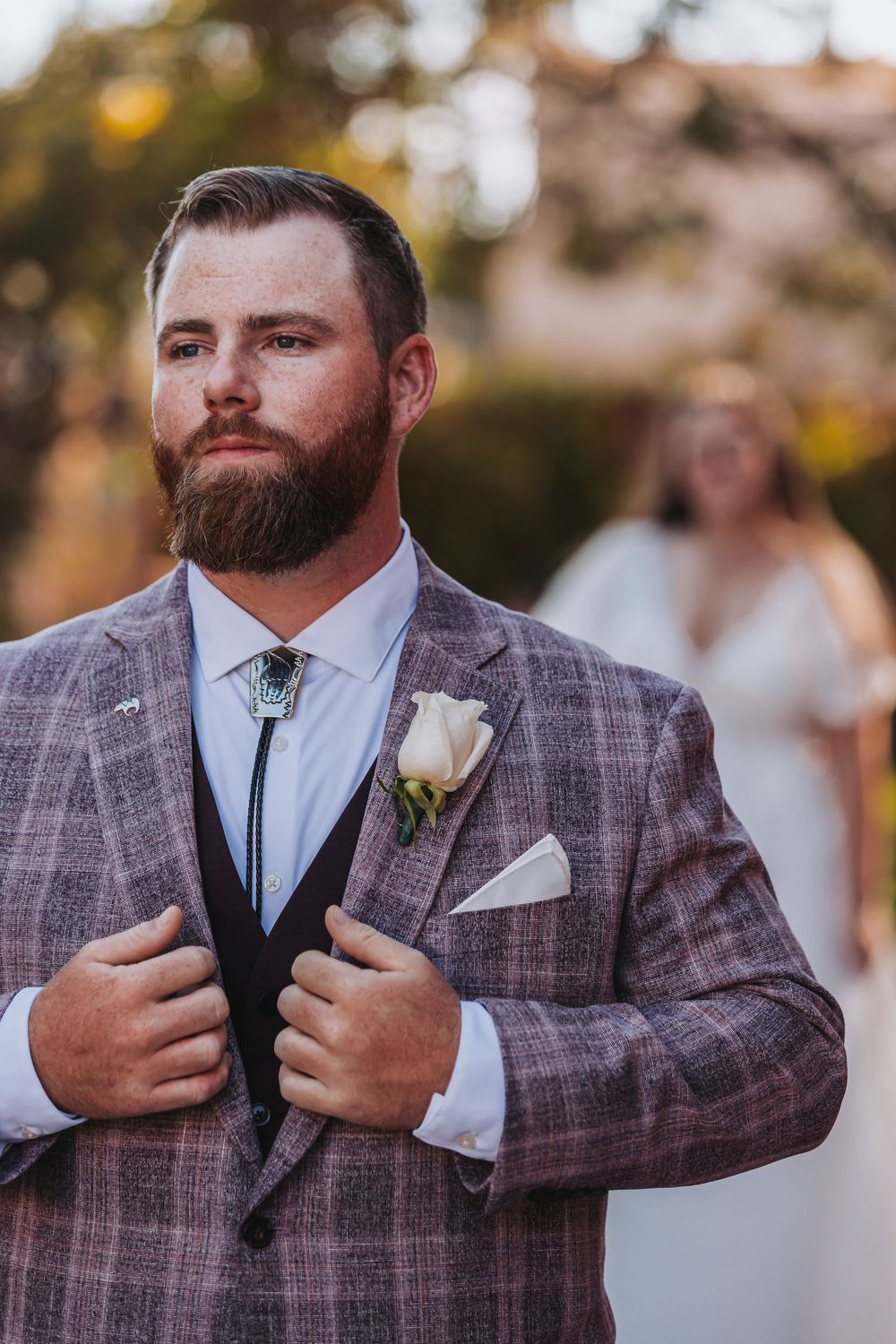 A groom in a plaid gray suit jacket with a white boutonniere adjusts his lapels during an outdoor wedding ceremony.