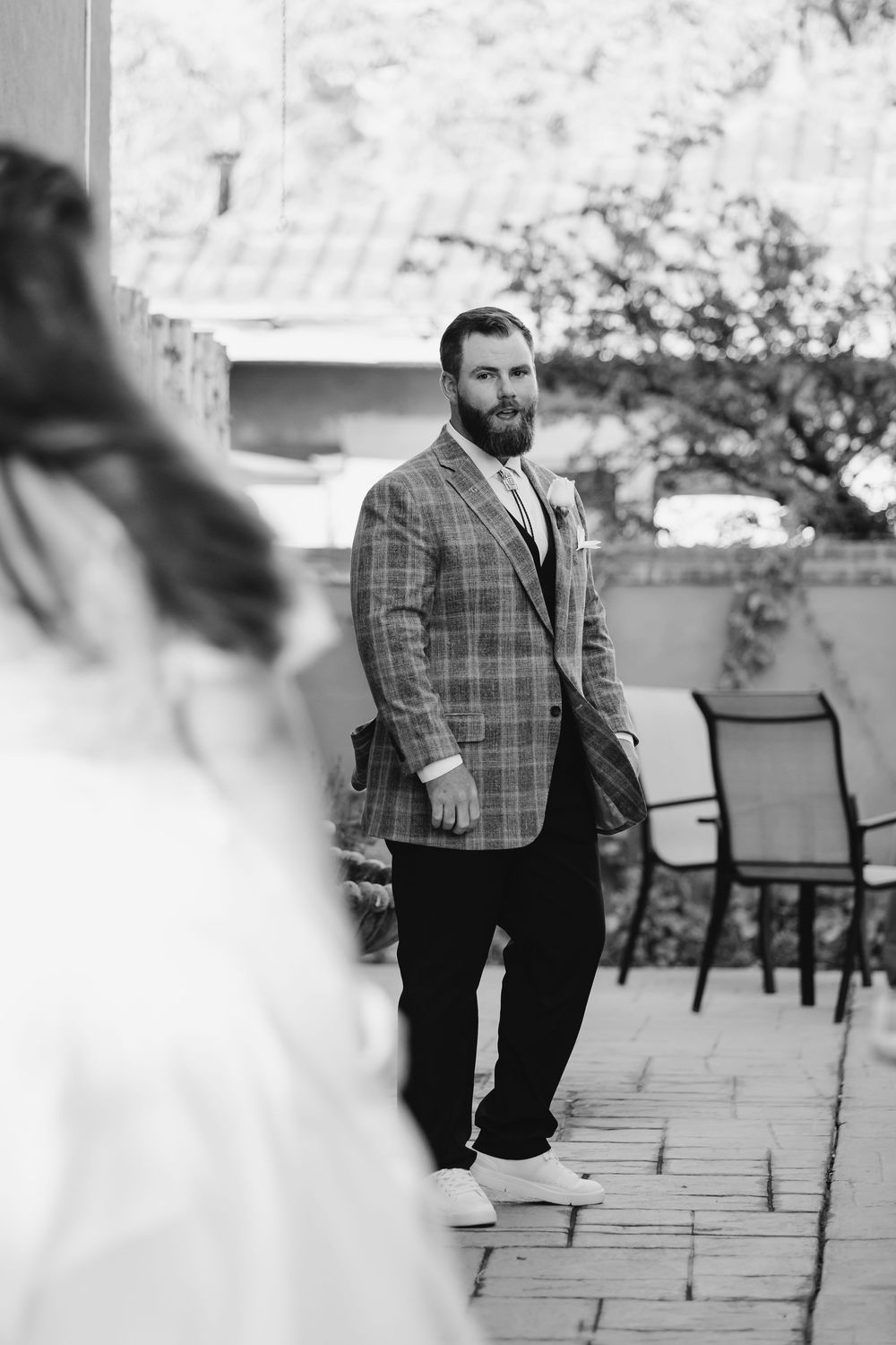 A person wearing a plaid suit jacket stands on a stone patio near outdoor chairs in black and white photography.