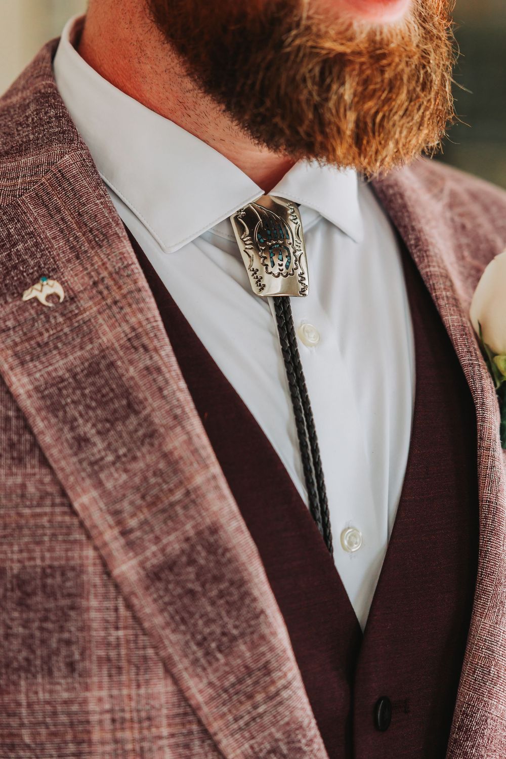 Close-up details of a brown tweed suit jacket with white dress shirt and black bolo tie against blurred background.
