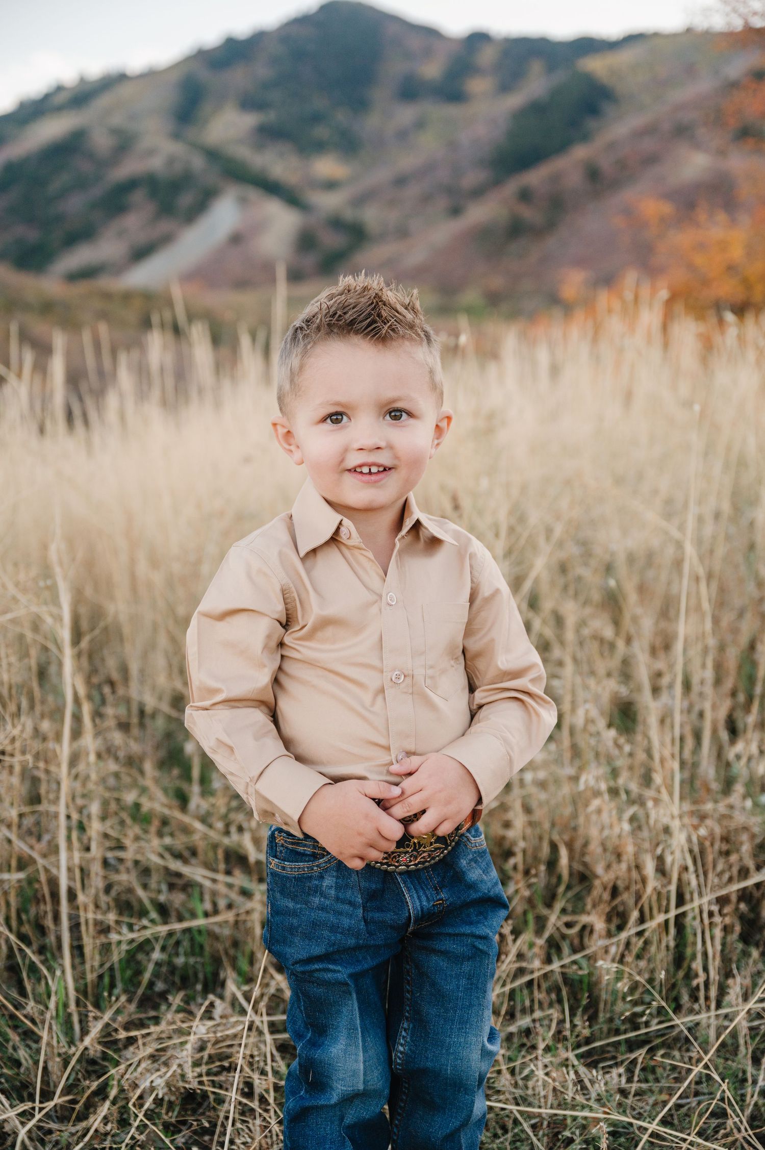 A young child stands in a golden autumn field wearing a beige button-down shirt and jeans with mountains in background.