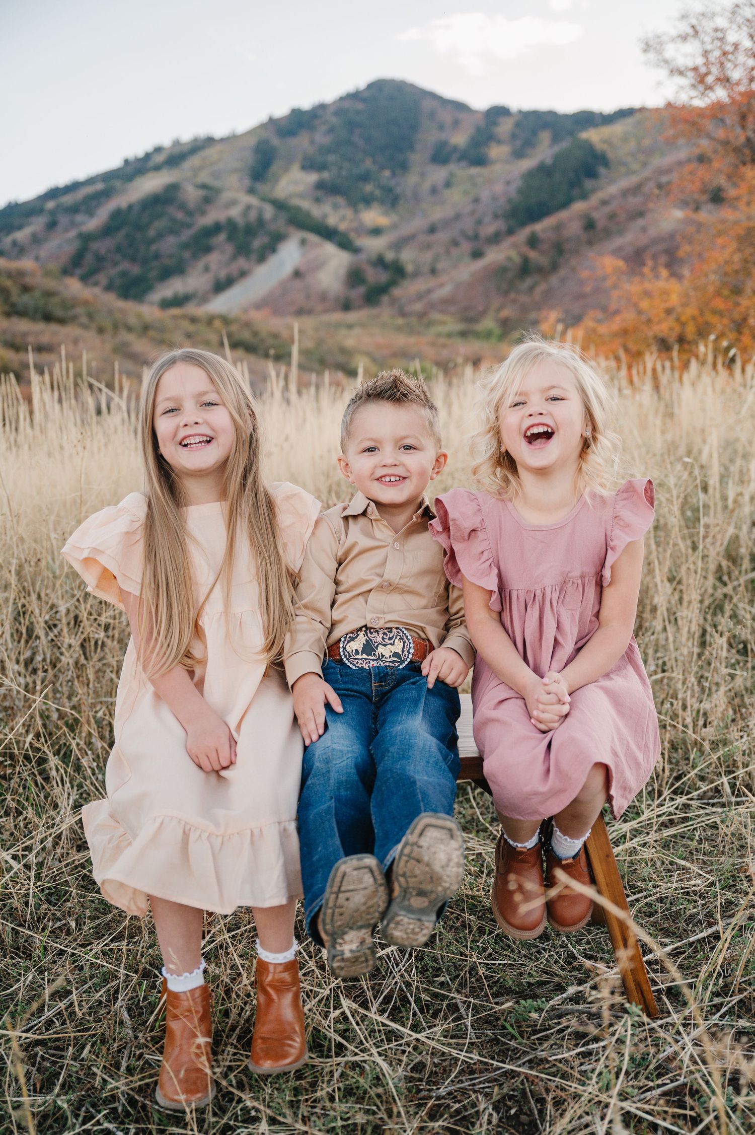 Children sitting together in tall grass laughing and playing during golden hour with mountains in background.