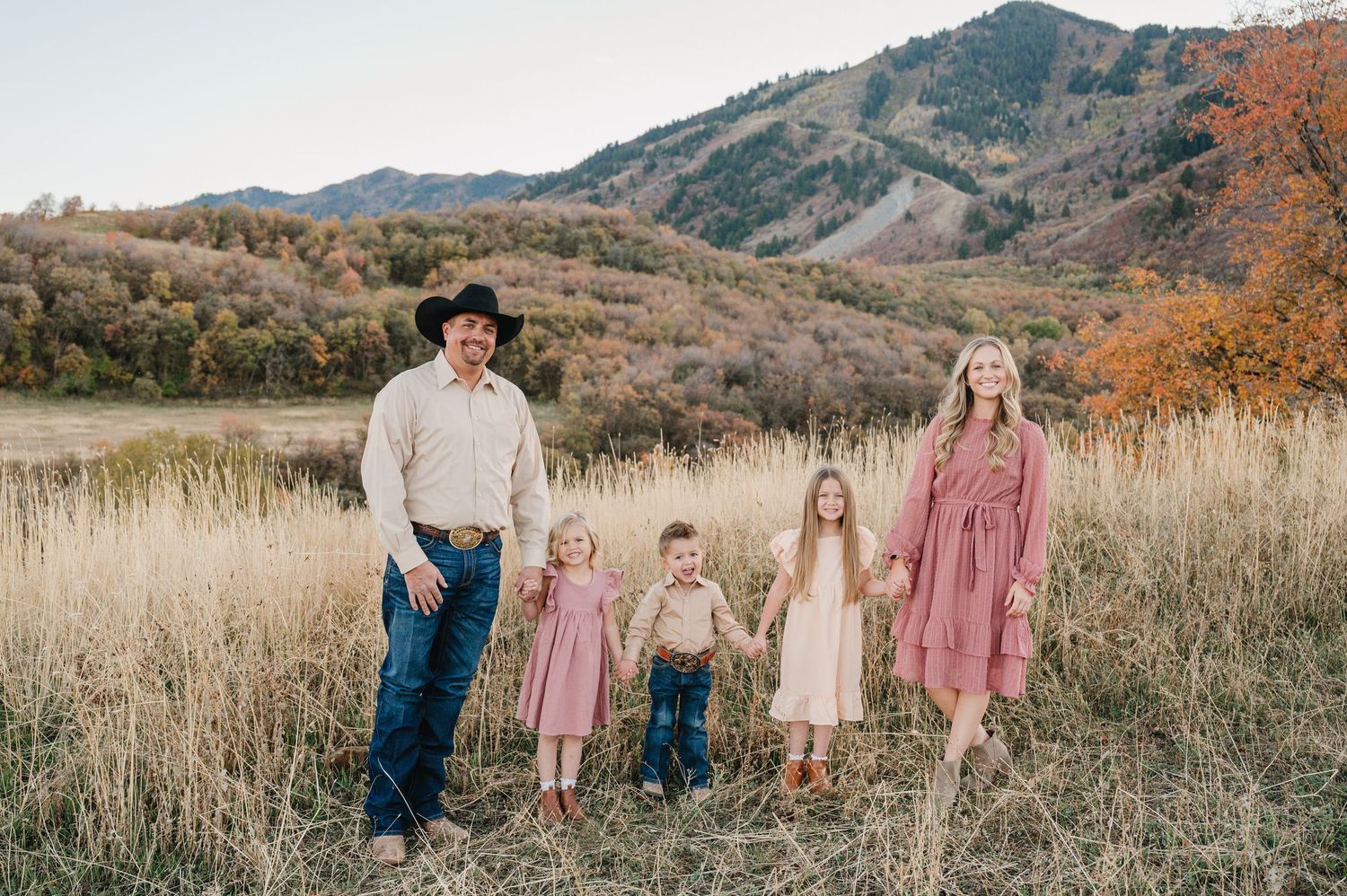 A family in coordinating outfits stands together in a golden field with mountains in the background during autumn.