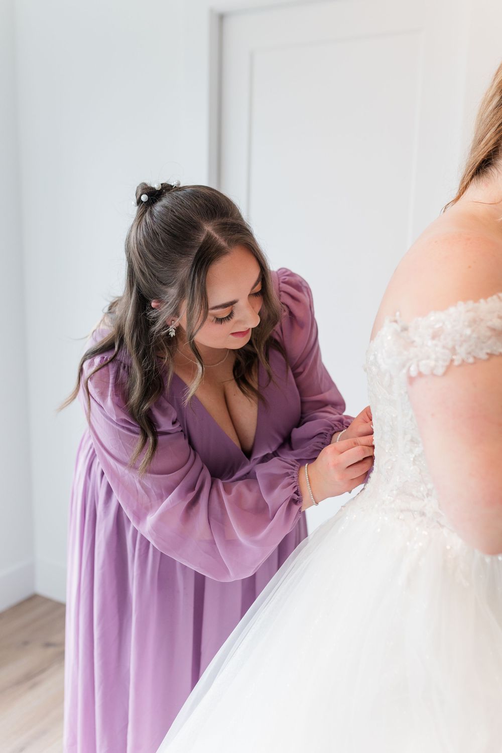 A bridesmaid in a lavender dress helps adjust the bride's white wedding gown in a sunlit room.