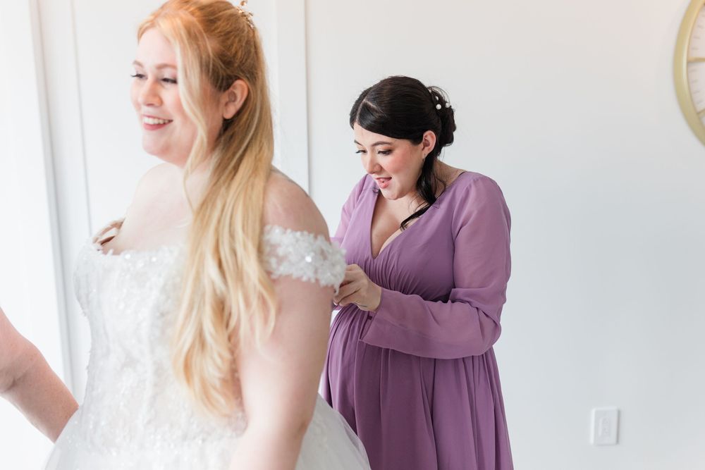 Bride getting ready with assistance from someone in a purple dress during wedding preparations.