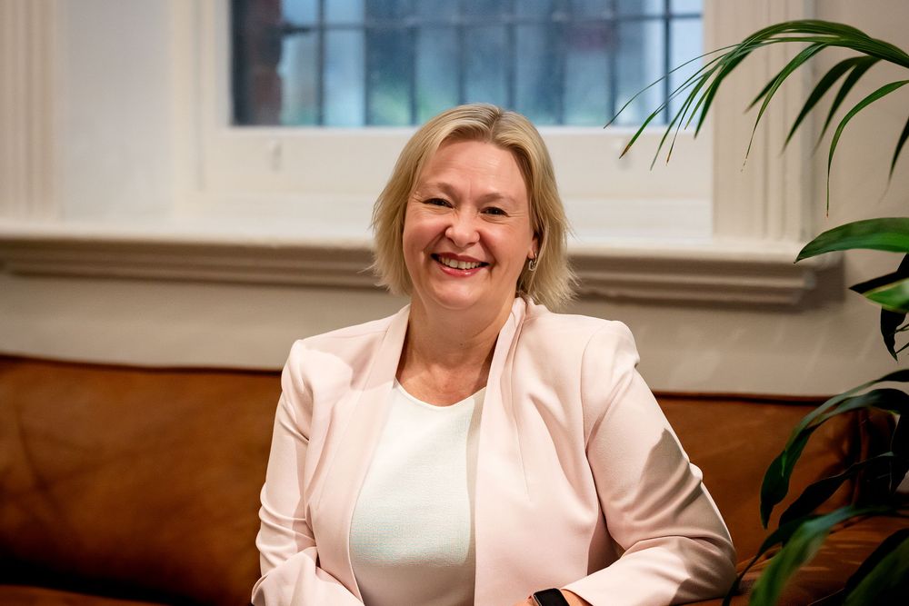 Smiling blonde woman in a light pink blazer seated on a leather sofa near a window and green plant.