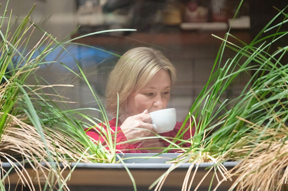 Blonde woman sipping coffee from a white cup, framed by tall green grass plants in a cozy cafe setting.
